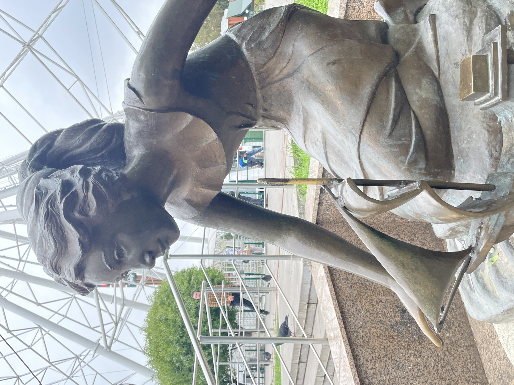 This is a medium shot, eye-level photograph of a bronze sculpture of a young girl in Kirov, Russia. The sculpture depicts the girl kneeling and holding a small sailboat. She is wearing a simple dress and her hair is pulled back in a ponytail. In the background, a Ferris wheel is visible, suggesting the sculpture is located in an amusement park or recreational area. There are also people walking in the background, and pigeons are present on the steps and railings near the sculpture. The sculpture is made of weathered bronze and appears to be placed on a stone pedestal. The overall lighting suggests an overcast day.