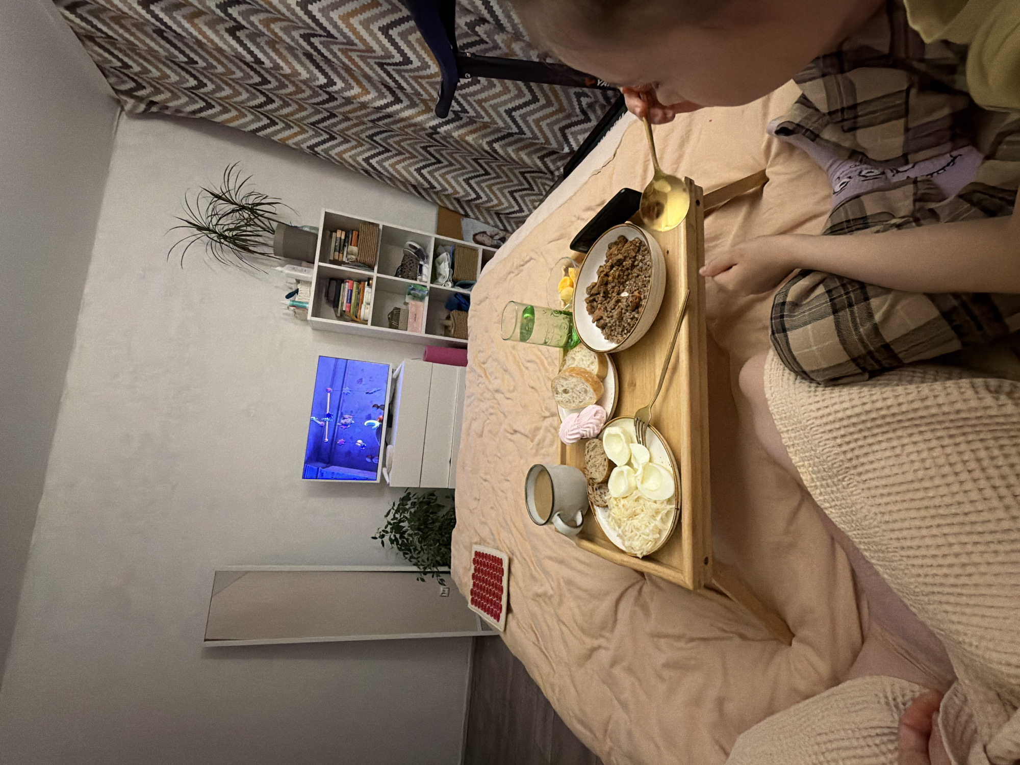A person is in bed in a room in Perm, Russia, eating breakfast from a wooden tray. The tray holds a bowl of food, a glass of juice, a plate with bread and marshmallows, a plate with eggs and shredded cheese, and a mug of coffee. The person is using a gold-colored spoon to eat. A TV screen shows an aquarium scene. A bookshelf, dresser, and potted plant are visible in the room. A gray-and-white chevron-patterned curtain hangs in the background.