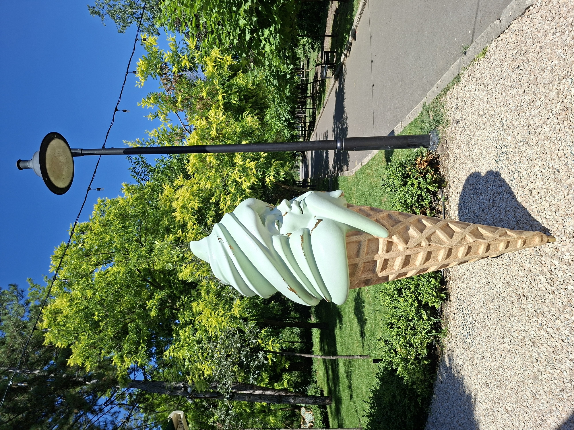This is a medium shot, taken outdoors on a sunny day, showing a large, mint-green ice cream cone sculpture with a face embedded in the top swirl of ice cream. The sculpture is made of a shiny material and appears to be melting slightly. The cone itself is a light brown, textured waffle cone.
The sculpture is positioned on a gravel path in the foreground. Behind the sculpture, to the left, is a grassy area with several trees, some with full green foliage and others with lighter, almost yellow-green leaves. A paved walkway runs to the right of the sculpture and extends into the background, lined with trees and park benches. A black lamppost with a large, circular light fixture stands to the right of the sculpture, and string lights are visible spanning between the lamppost and another unseen pole in the distance.
The scene appears to be a park or public garden area. The overall impression is one of a cheerful, somewhat whimsical outdoor space. The location of this sculpture is identified as being in the Chișinău, Moldova.