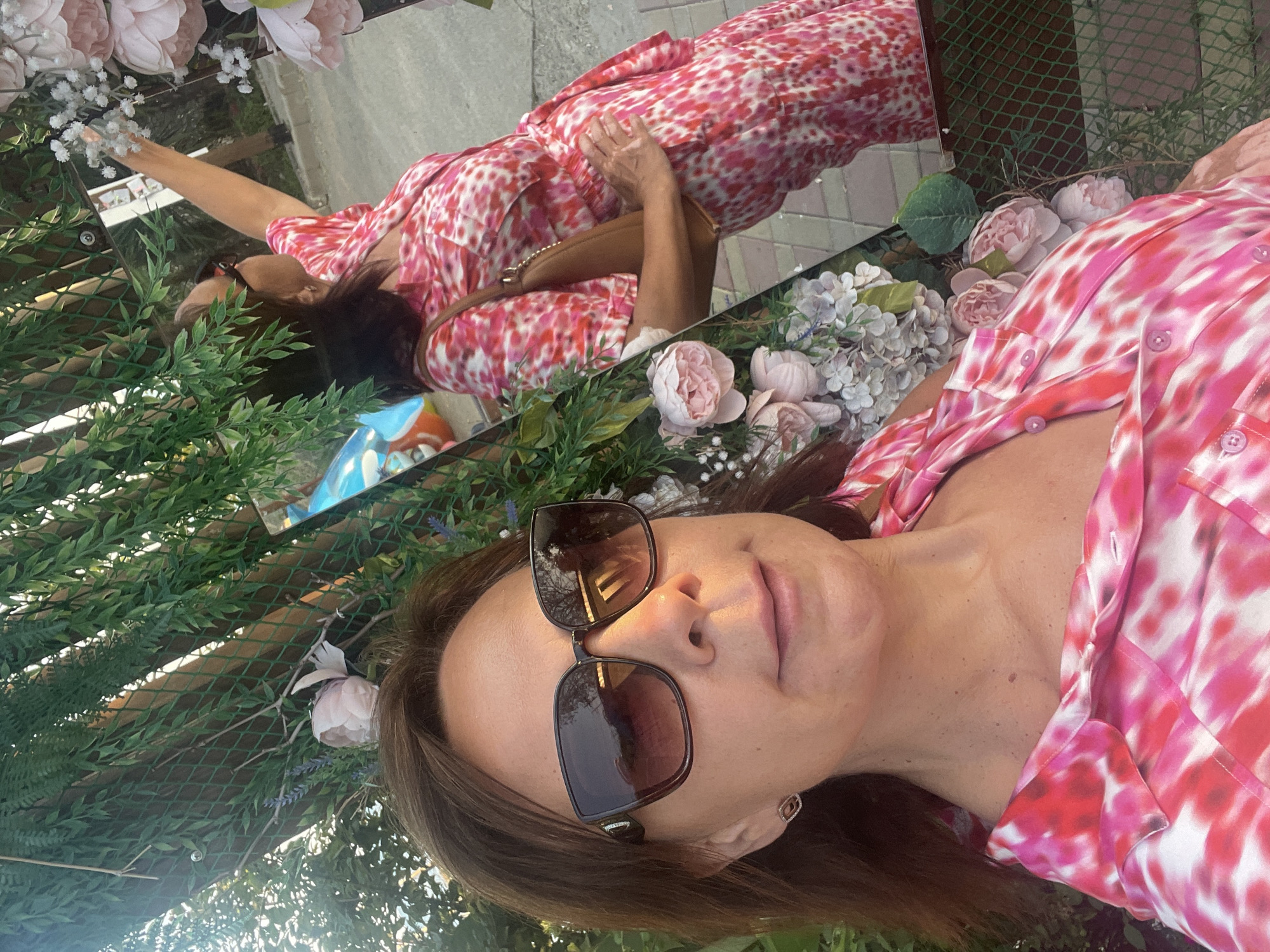 A woman, wearing a pink and white tie-dye button-down shirt and large sunglasses, takes a selfie. She is standing in front of a mirror, which reflects her and a pink floral arrangement. In the reflection, she is reaching out to touch the flowers. She is wearing a brown crossbody bag. The background suggests an outdoor setting with greenery and what appears to be a wooden fence. The image seems to be taken in the city of Sochi, Russia, possibly at an outdoor cafe or garden.
