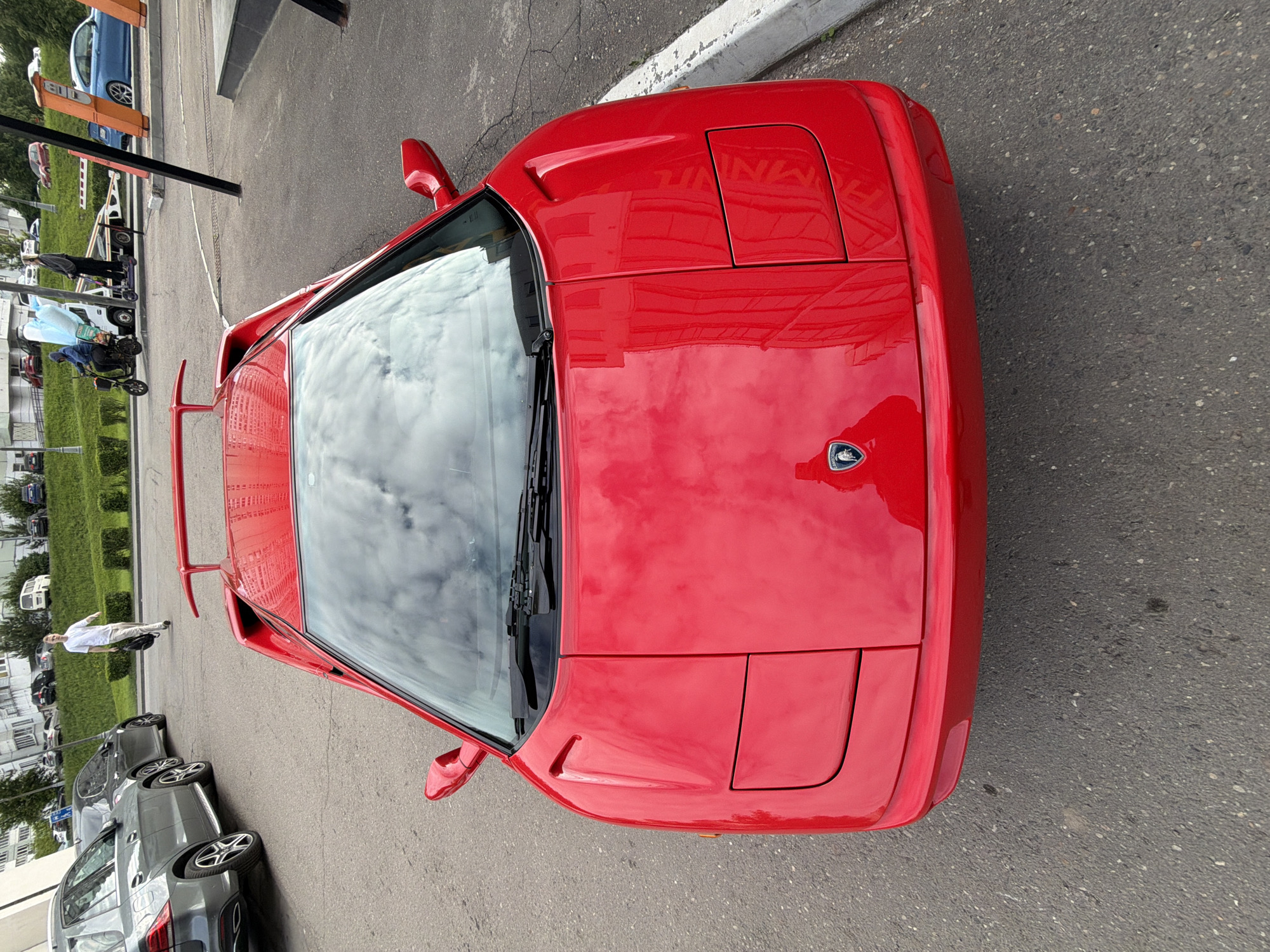 The image captures a vibrant red Lamborghini sports car, likely a Diablo model, positioned prominently on an asphalt surface, viewed from an elevated, slightly angled perspective. The car's sleek design is evident, featuring closed pop-up headlights and a distinctive rear wing. Its highly reflective red paintwork shows clear reflections of clouds and architectural structures from the surroundings. The iconic Lamborghini emblem is visible on the center of the front hood.
In the background, an urban environment unfolds. To the left, a row of dark gray cars is parked, and a blue sedan is visible further down a street or lane, partially obscured by a black barrier post. Several individuals are present in the scene: a person in a white shirt is seen standing further back on the left, while others are visible near scooters and walking along a paved area or sidewalk. Green hedges and bushes line the distant background, adding a touch of greenery to the urban setting.
The scene takes place during the daytime, under what appears to be an overcast or partly cloudy sky, as suggested by the reflections on the car's surfaces and windshield. The ground is paved with asphalt, marked by a white curb line. The overall impression is a bustling urban setting, with the striking sports car as the focal point. The visible text is limited to the Lamborghini emblem on the car's hood.