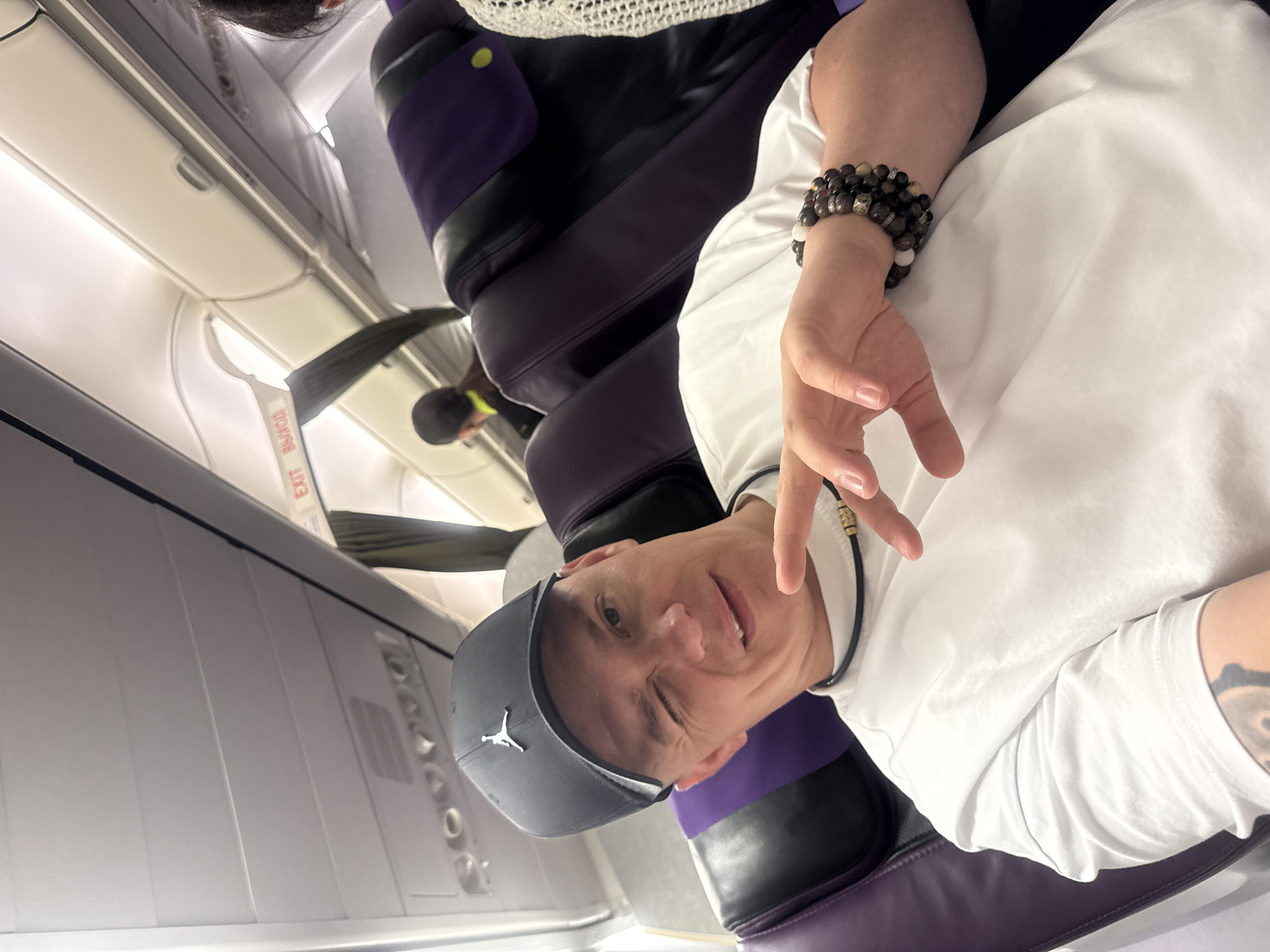 The image shows a man on an airplane. He is wearing a white t-shirt, a grey baseball cap with a Jumpman logo, and a beaded bracelet on his right wrist. He is winking and making a peace sign with his fingers. The airplane cabin is visible, with purple and black seats. An "EXIT" sign is also visible. The lighting suggests it is daytime. The man appears to be in a good mood, possibly on vacation or traveling for leisure. The setting is an airplane, suggesting travel. No specific time of day or weather is discernible.