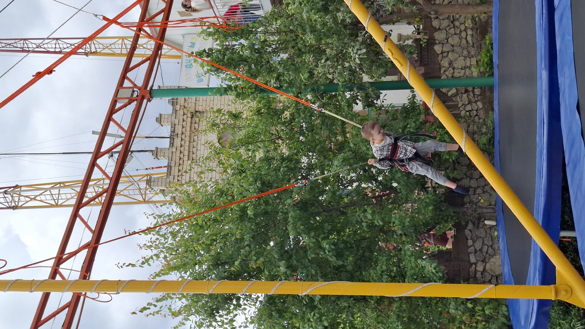The image shows a child wearing a harness attached to bungee cords suspended from a yellow and red metal frame above a trampoline. The child is mid-air, appearing to jump or bounce. The scene is set outdoors with trees and a brick building visible in the background. The location appears to be a commercial or recreational area in an unidentified city. The sky is overcast.