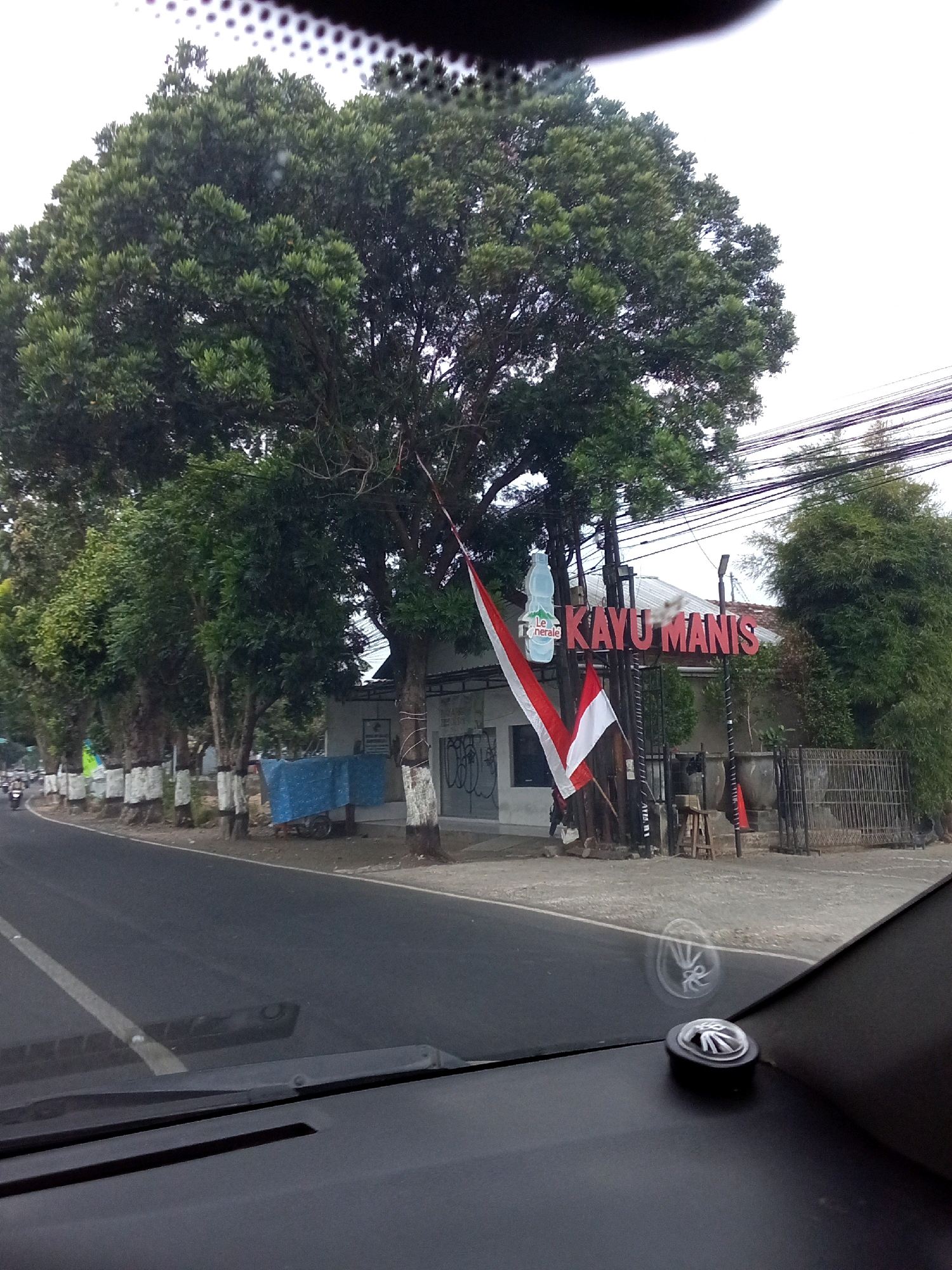 The image depicts a street scene in Ngasem, Indonesia. In the foreground, the dashboard and windshield of a vehicle are visible, with a small black accessory mounted on the dashboard. The scene outside the vehicle shows a paved road with a white line marking the lane. Along the side of the road, several trees with white-banded trunks are planted. To the right of the road, a building with a white exterior is present. Above the building, a large, leafy tree dominates the upper left portion of the image, with power lines strung across the sky. A sign with red lettering reads "KAYU MANIS," and a white bottle-shaped logo with green lettering "Le Minerale" is also visible. Two Indonesian flags, one red and white and the other all white, are hung on a pole near the sign. A small wooden stool and some potted plants are also present. A blue tarp covers an object on a cart. In the background, more trees and vegetation are visible. The sky is overcast.