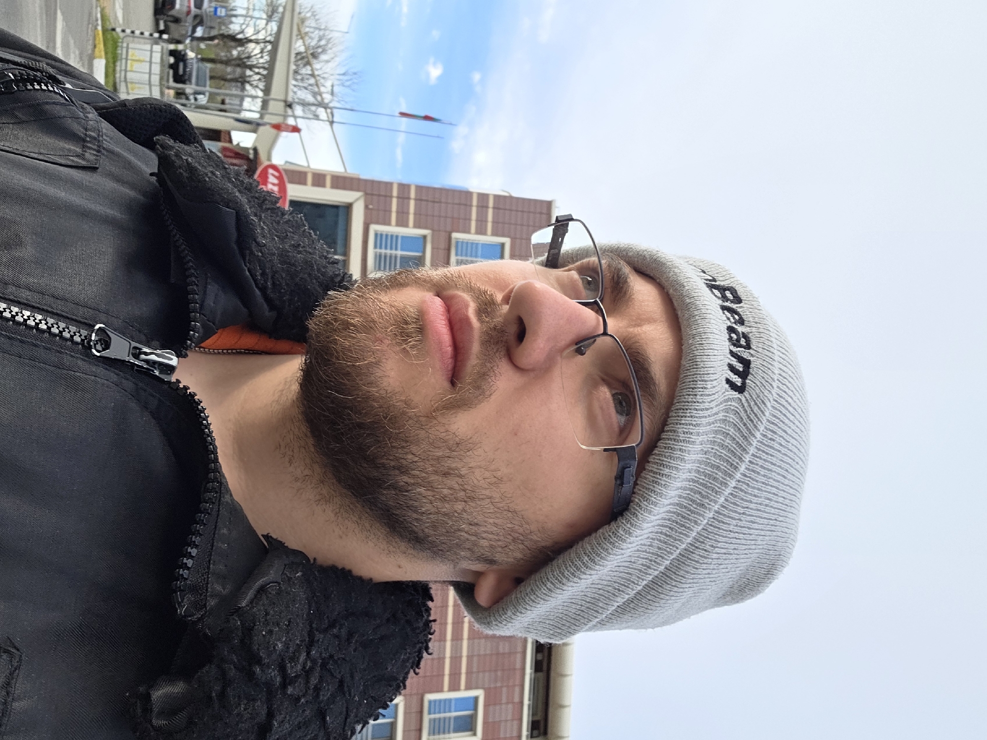 A selfie of a man wearing a gray beanie with the word "beam" written on it and glasses. He is wearing a black jacket with a fuzzy collar and a visible zipper. The background features a brick building with windows and a blue sky with some clouds. A street sign is also visible in the background. The location is likely in an urban setting. The information about Plovdiv, Bulgaria cannot be verified from the image.