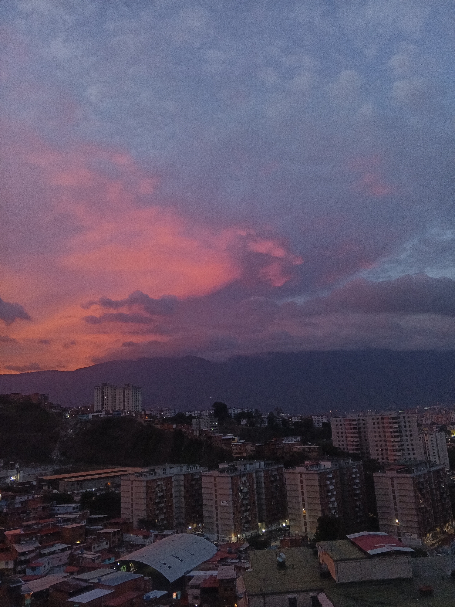 The image captures a dusk scene in Caracas, Venezuela, featuring a cityscape under a sky streaked with vibrant pink and gray clouds. The silhouette of a mountain range is visible in the background. Several tall apartment buildings dominate the midground, interspersed with smaller residential structures with red roofs. The lighting is dim, with pockets of artificial light emanating from some buildings. There are no people visible in the image.