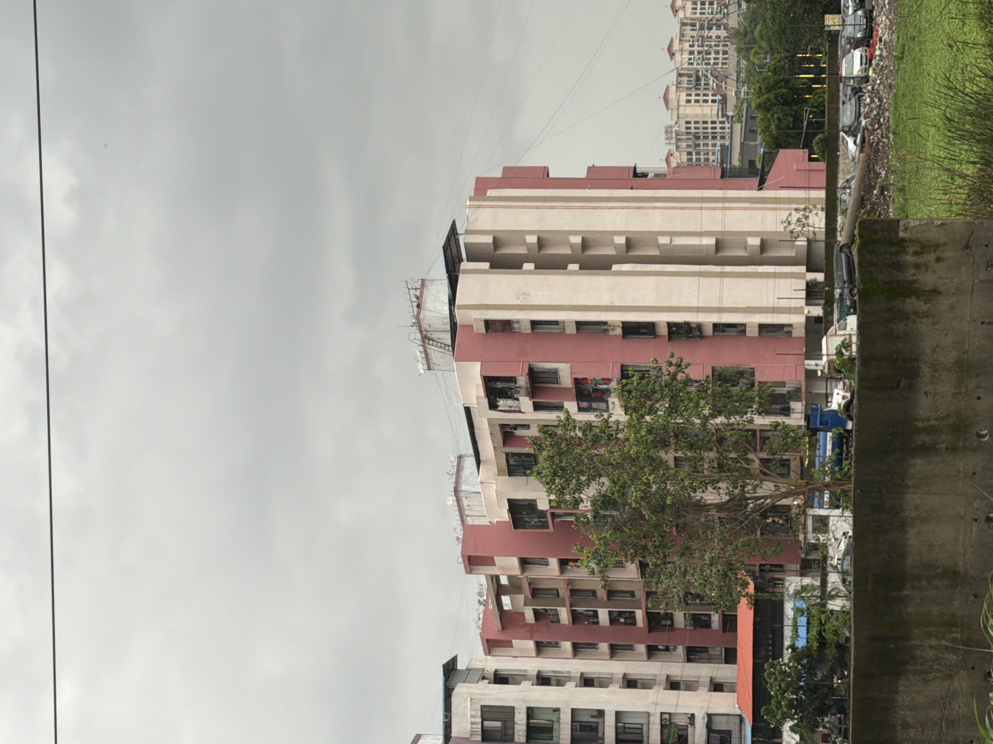 The image depicts a cloudy day in Vasai-Virar, India. In the foreground, there is a concrete wall with some greenery growing on its surface. Beyond the wall, a patch of overgrown grass and weeds is visible, with some debris scattered around.
Dominating the midground are several multi-story buildings. The building in the center is painted in sections of pink and cream, featuring numerous windows and balconies. To its left is a taller, predominantly cream-colored building with a lighter colored section on the far left, also featuring many windows. Behind these structures, other buildings with similar architectural styles are visible, including one with a red roof in the lower left.
Parked vehicles are present in front of the buildings, including a blue and white truck, several cars, and a white van. A tree with green foliage stands in front of the pink and cream building, partially obscuring its lower levels.
In the background, a dense cluster of residential buildings stretches across the horizon under the overcast sky. Power lines are visible crisscrossing the upper portion of the image. The overall scene suggests an urban or semi-urban environment characterized by residential architecture and overcast weather conditions.