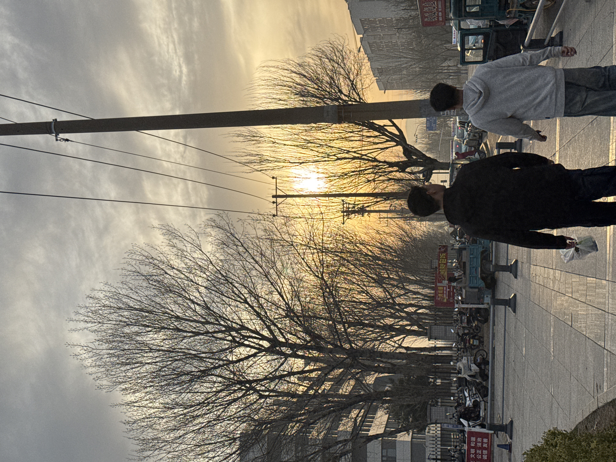 The image shows a street scene in Zhongtun, China, at sunset. A sidewalk lined with bare trees runs alongside the street. Two people are walking along the sidewalk, facing to the right. One person is wearing a grey hoodie, the other is wearing a black jacket. Numerous parked motorcycles and small vehicles are visible along the sidewalk. Signs with Chinese characters are posted in the background. Overhead power lines stretch across the sky, silhouetting the trees against the setting sun. Buildings are visible in the background on both sides of the street. The sky is a mix of cloudy grey and golden hues from the sunset.