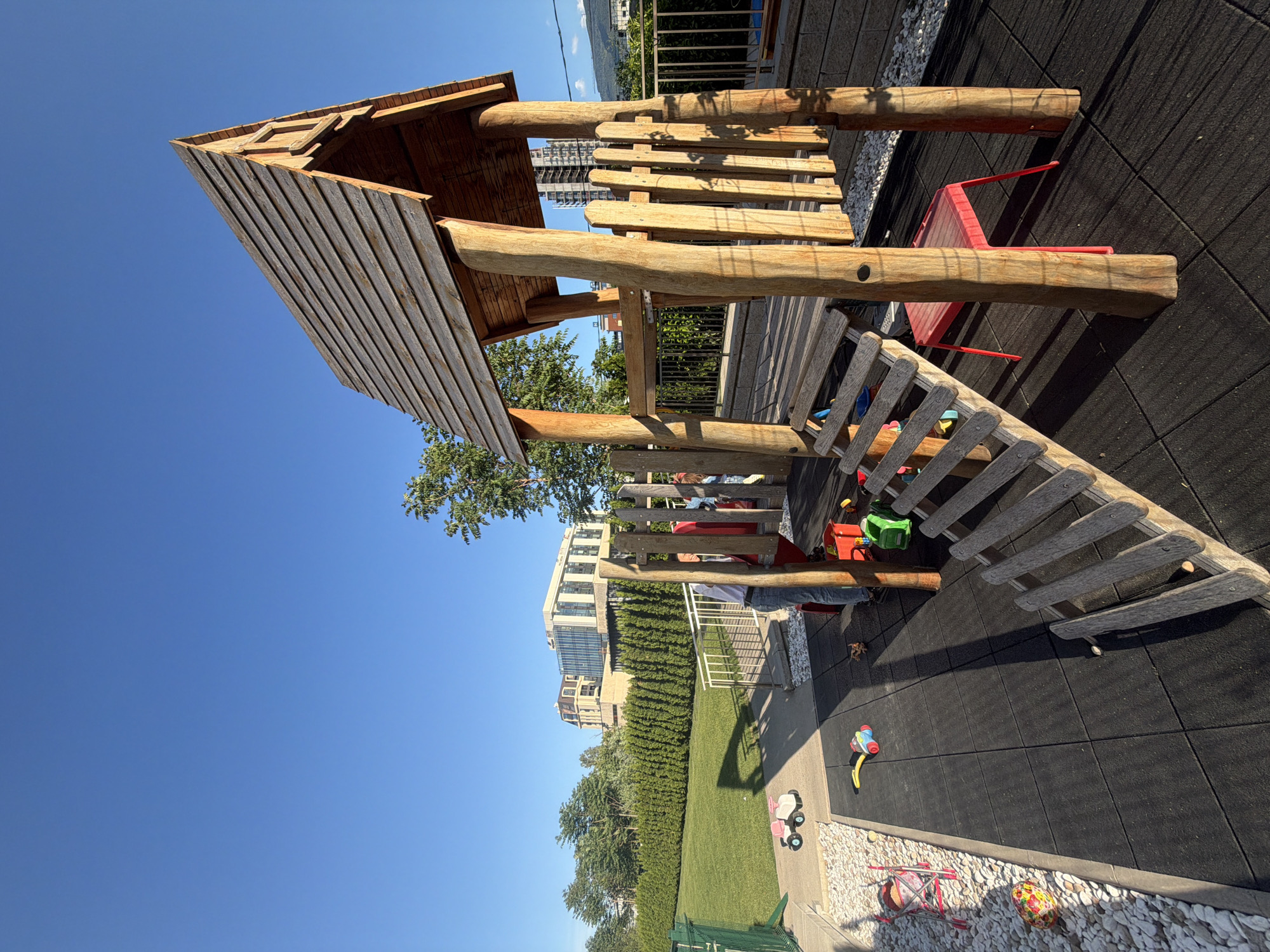 The image depicts a children's playground in Sofia, Bulgaria, featuring a wooden play structure with a roof, ladder, and seating area. The playground is located outdoors, with a backdrop of buildings and trees visible in the distance. The ground surface is covered with black rubber tiles. Scattered around the play structure are various children's toys, including a tricycle, a toy car, and other smaller playthings. A red chair is also visible near the wooden structure. The sky is clear and blue, indicating a sunny day. The play structure appears to be made of unfinished wood logs and planks.
