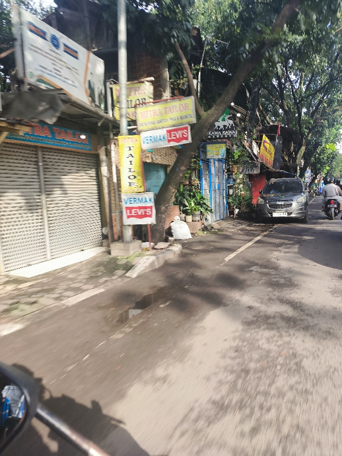 The image shows a street in Bandung, Indonesia (latitude: -6.901, longitude: 107.647) with a closed garage door and several shops on the left side of the frame. There are multiple signs in Indonesian and English for tailors and a bakery. A large tree trunk is visible in the center of the frame, with a white bag lying beneath it. There is a gray car parked on the right side of the street, facing the camera. A motorcycle is visible at the bottom of the frame, seemingly taken from the perspective of the rider. The street is asphalt and the day is sunny. The image is taken from a moving vehicle.