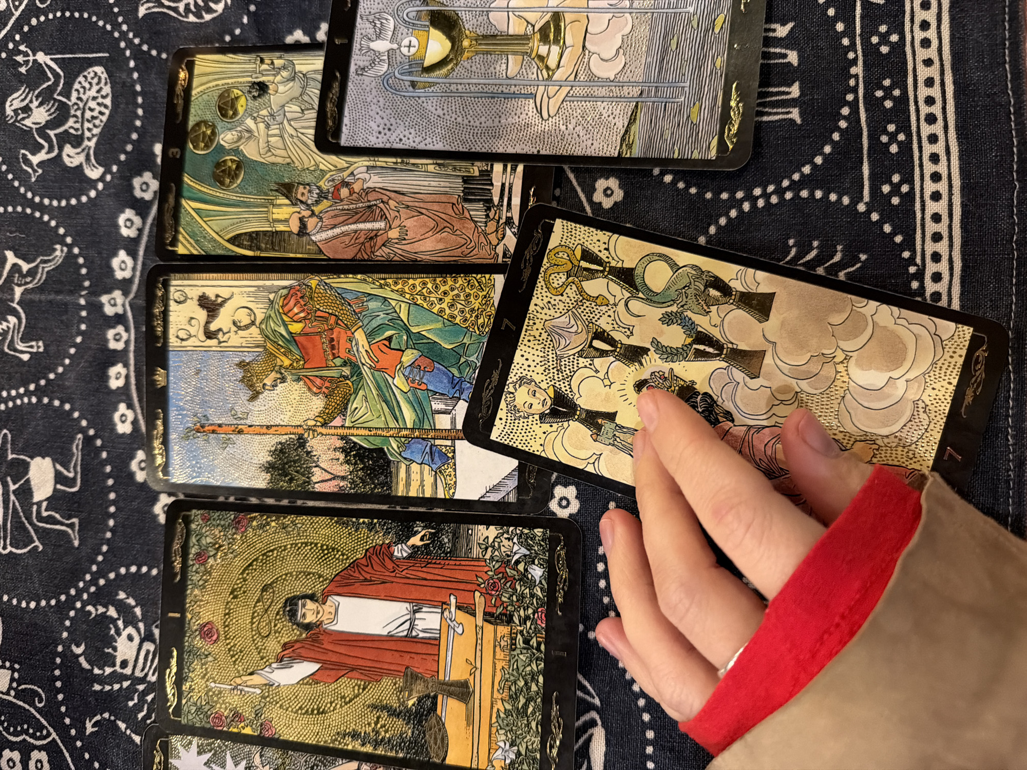 This close-up shot captures a hand interacting with a spread of several tarot cards laid out on a dark blue patterned cloth. The setting is likely indoors, in Tel-Aviv, Israel, although no specific environmental cues confirm the location.
The subject's left hand, with a silver ring on the ring finger and a visible red lining of a sleeve, gently rests on one of the cards, possibly in the midst of a tarot reading or arrangement.
Several tarot cards are visible:
- At the top center, a card depicting a golden chalice held by hands, possibly the Two of Cups.
- To its left, the Three of Pentacles is shown, featuring three figures engaged in work or consultation with three golden pentacles.
- Below the Three of Pentacles, a card depicting a robed figure holding a staff, reminiscent of a Court card from the Wands suit (e.g., King or Page of Wands).
- Partially obscured by the hand, the Seven of Wands is visible, showing a person defending against multiple wands. The number "7" is clearly seen on this card.
- At the bottom center, The Magician card (Major Arcana I) is displayed, featuring a young man with a red robe, holding a wand, with various suit symbols (cup, sword, pentacle) on a table before him, and roses above.
The cards feature a detailed, illustrative style with gold metallic accents and textured backgrounds. The dark blue cloth beneath them has white intricate patterns, including dotted circles, floral motifs, and stylized figures. The overall scene suggests a moment of contemplation or a ritualistic act involving the divination tools.
