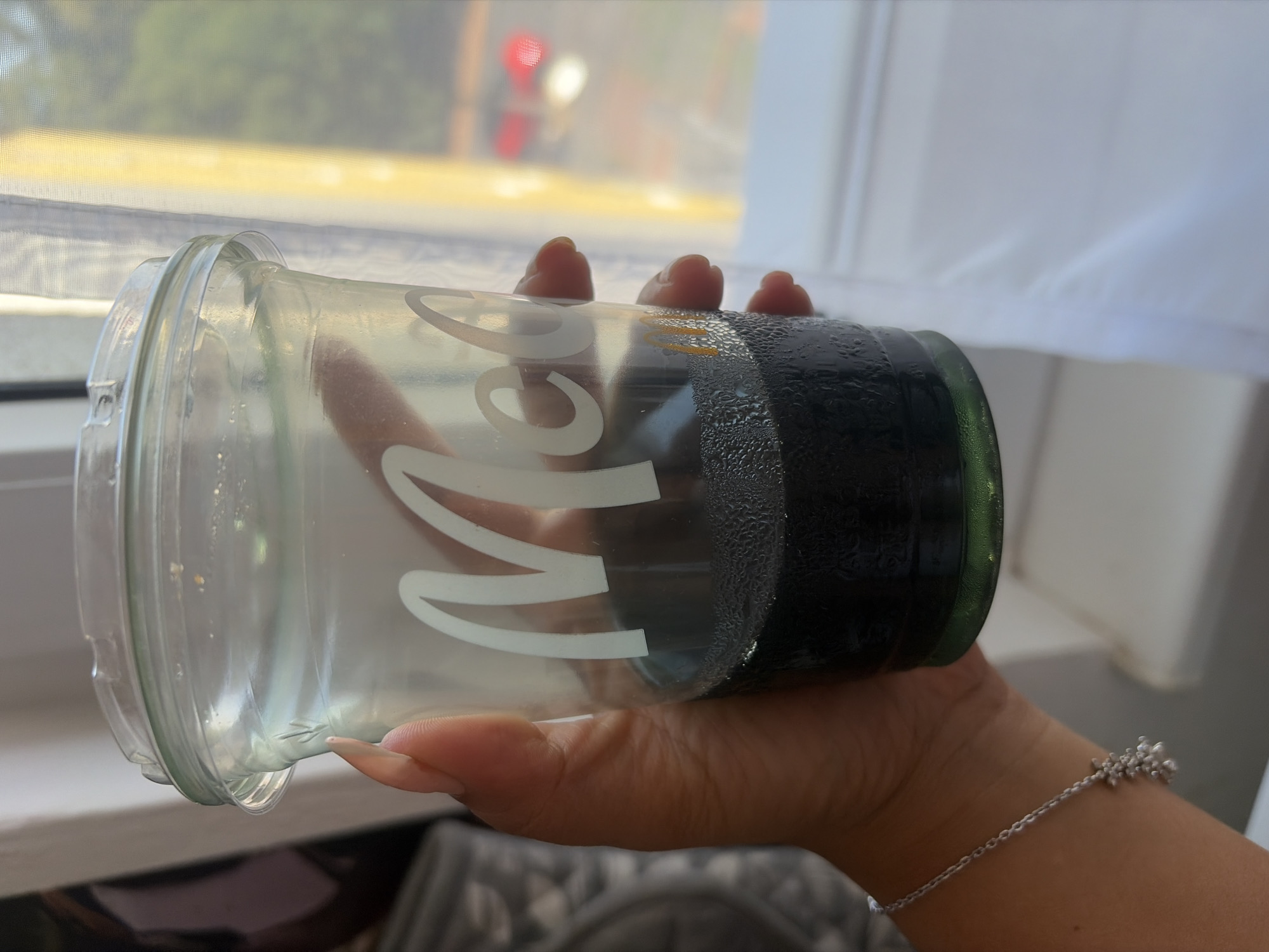 A hand with painted nails holds a clear plastic cup with a condensation-covered dark liquid. The cup has a white "Mc" logo on it. The background shows a window with sheer white curtains and a blurred outdoor scene with a red car.