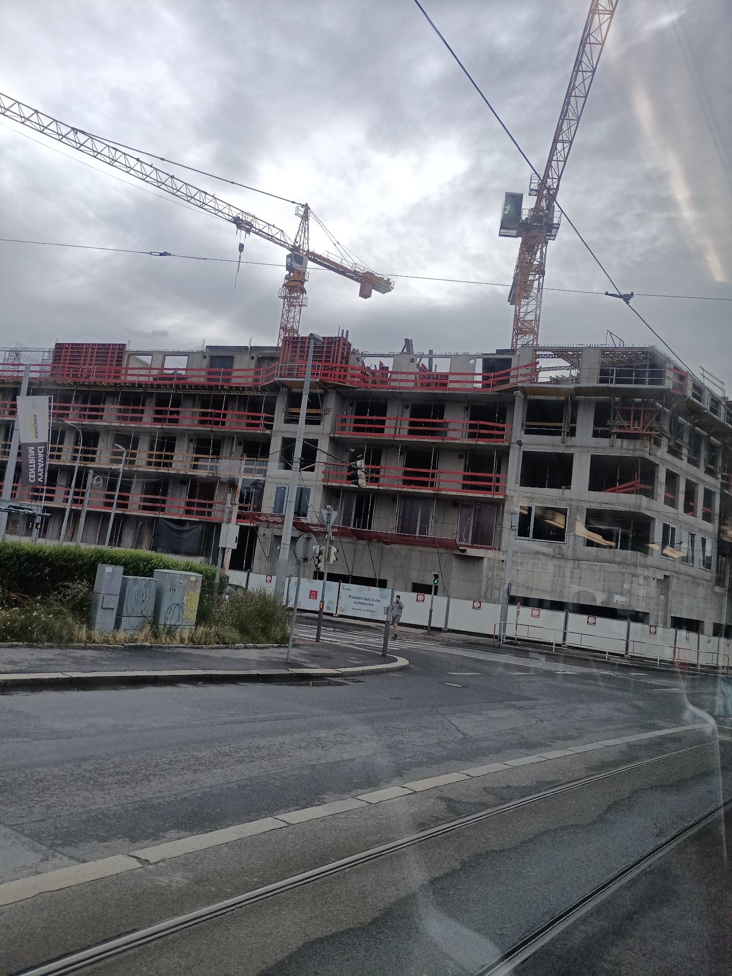 This image shows a large construction site with a multi-story building under construction. Two tower cranes are visible, one on the left and another taller one on the right, dominating the skyline. The building itself is made of concrete and steel, with red safety railings lining the open floors. Several windows are visible, some with glass and others empty frames. The scene appears to be a typical day at a construction site, with no people clearly identifiable, though there are indications of activity.
The weather appears to be overcast and cloudy, suggesting a potentially humid or rainy day, as evidenced by the wet road surface in the foreground. The time of day is indeterminate due to the lack of direct sunlight or strong shadows.
In the foreground, a wet asphalt road with tram tracks runs across the bottom of the frame. On the left side, there is a patch of overgrown greenery and some electrical boxes, one of which has graffiti. A white fence surrounds the construction site, with various signs. One sign on the left, partially obscured, has text that is difficult to read due to the angle and resolution, but it appears to be in a European language, possibly Czech given the location context of Prague. Another sign on the right has a circular logo with a stylized "C" inside and text below it, also in a European language.
The overall impression is one of ongoing development and urban transformation in a modern city. The presence of tram tracks and the architecture hint at an urban European setting, specifically Prague, Czechia.