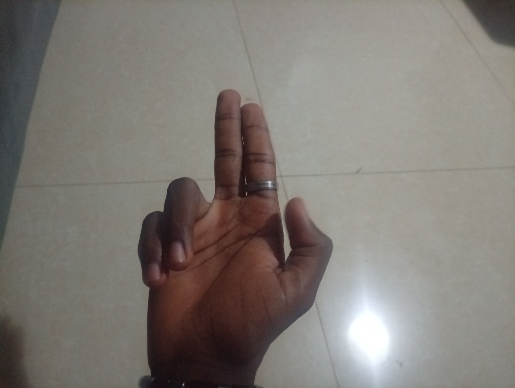 A medium, eye-level shot shows a brown-skinned hand extended upward. The middle and index fingers are raised together, a silver ring is on the middle finger, and the other fingers are curled downward toward the palm. A dark-colored bracelet is seen on the wrist. The background is a beige tiled surface with a glare of light on the lower right. The location is Unknown, Unknown.