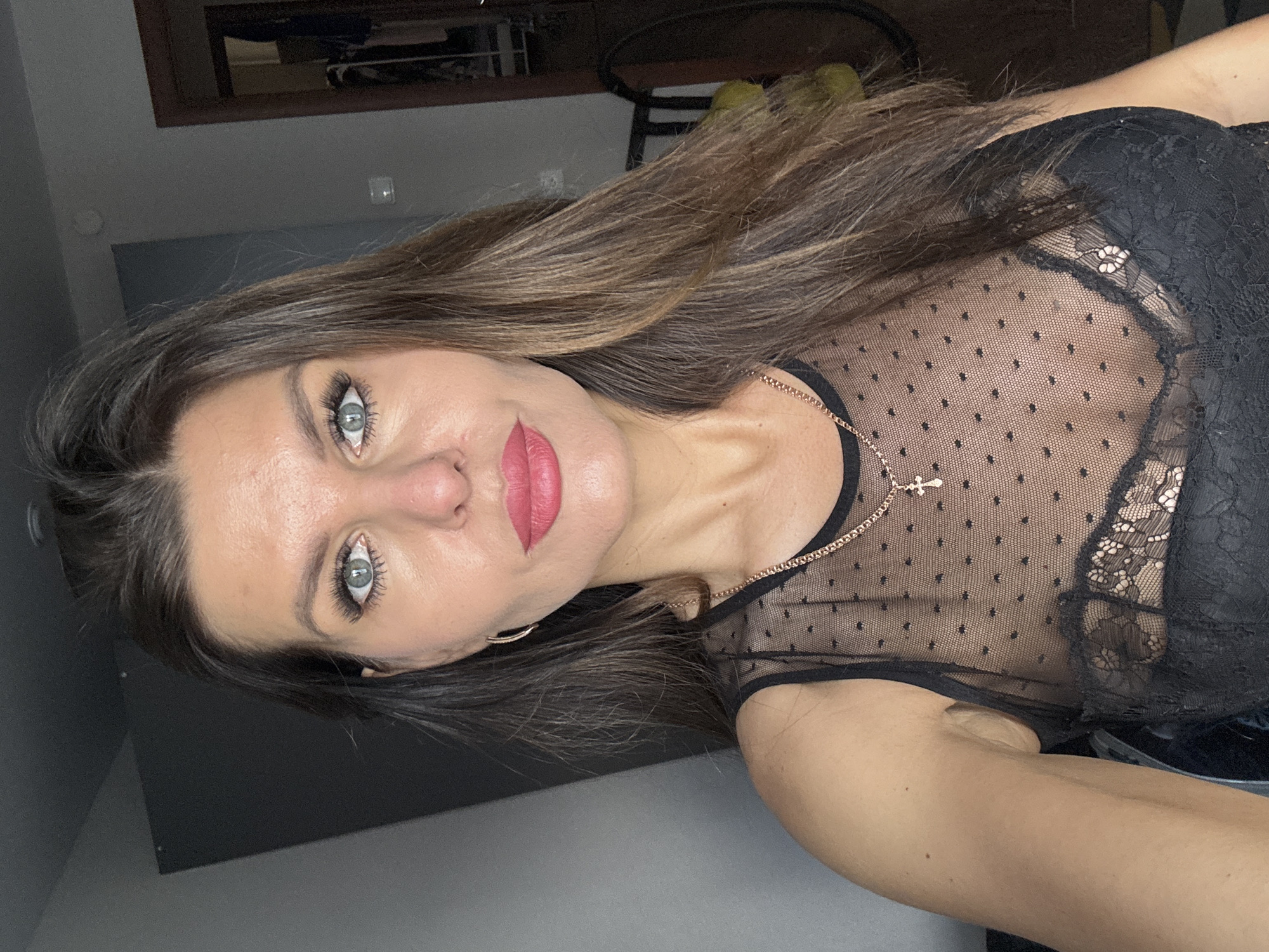 A woman with long brown hair and light blue eyes is pictured in a selfie. She wears a black, sheer top with a small gold necklace with a cross pendant. The background appears to be an indoor space with a dark wall and a glimpse of furniture. The image provides no details that would associate it with the city of Chaikovsky, Russia.