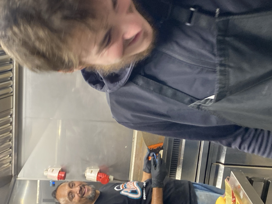 Two men are visible in a commercial kitchen. The man closer to the camera, partially cropped, has light hair and a beard, wearing a black hooded sweatshirt and apron. The other man is wearing a black t-shirt and black gloves. He smiles and holds a small orange and black tool. Behind them, are stainless steel walls and equipment, with visible electrical outlets and ventilation. The scene is brightly lit, suggesting a functional workspace.