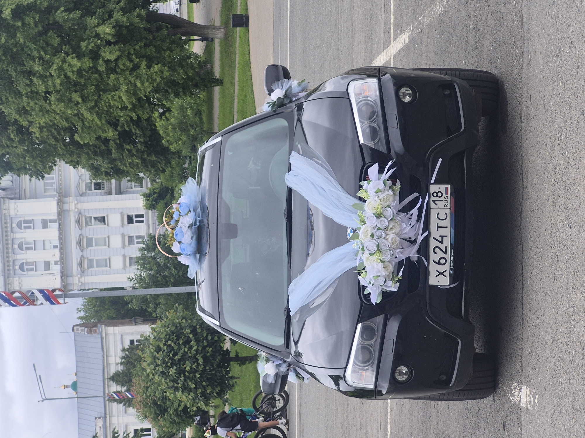 A black BMW X3 SUV, decorated for a wedding, is centrally parked on asphalt pavement. The vehicle features an arrangement of white and light blue artificial flowers with light blue sheer fabric ribbons draped over the front hood. A smaller floral arrangement, also in white and light blue, sits on the roof, topped with two interconnected golden rings. Small clusters of similar flowers are affixed to the side mirrors. The front license plate displays "X624TC 18 RUS".
In the background, under an overcast sky, the urban environment of Sarapul, Russia, is visible. To the far left, two partially obscured individuals on bicycles are present, with one wearing a helmet and backpack. Behind them, green foliage partially conceals a building with a grey roof and a prominent green onion dome, suggesting a religious structure. A smaller flag with Russian national colors is faintly visible near this building. Towards the center-background, a tall, slender pole displays a spiraling ribbon in the white, blue, and red colors of the Russian flag. Adjacent to the pole stands a stately, light-colored, multi-story building with numerous windows and classical architectural details. To the right, large mature green trees are present, along with a dark cylindrical waste bin. The ground is asphalt with faded white parking lines visible on the right.