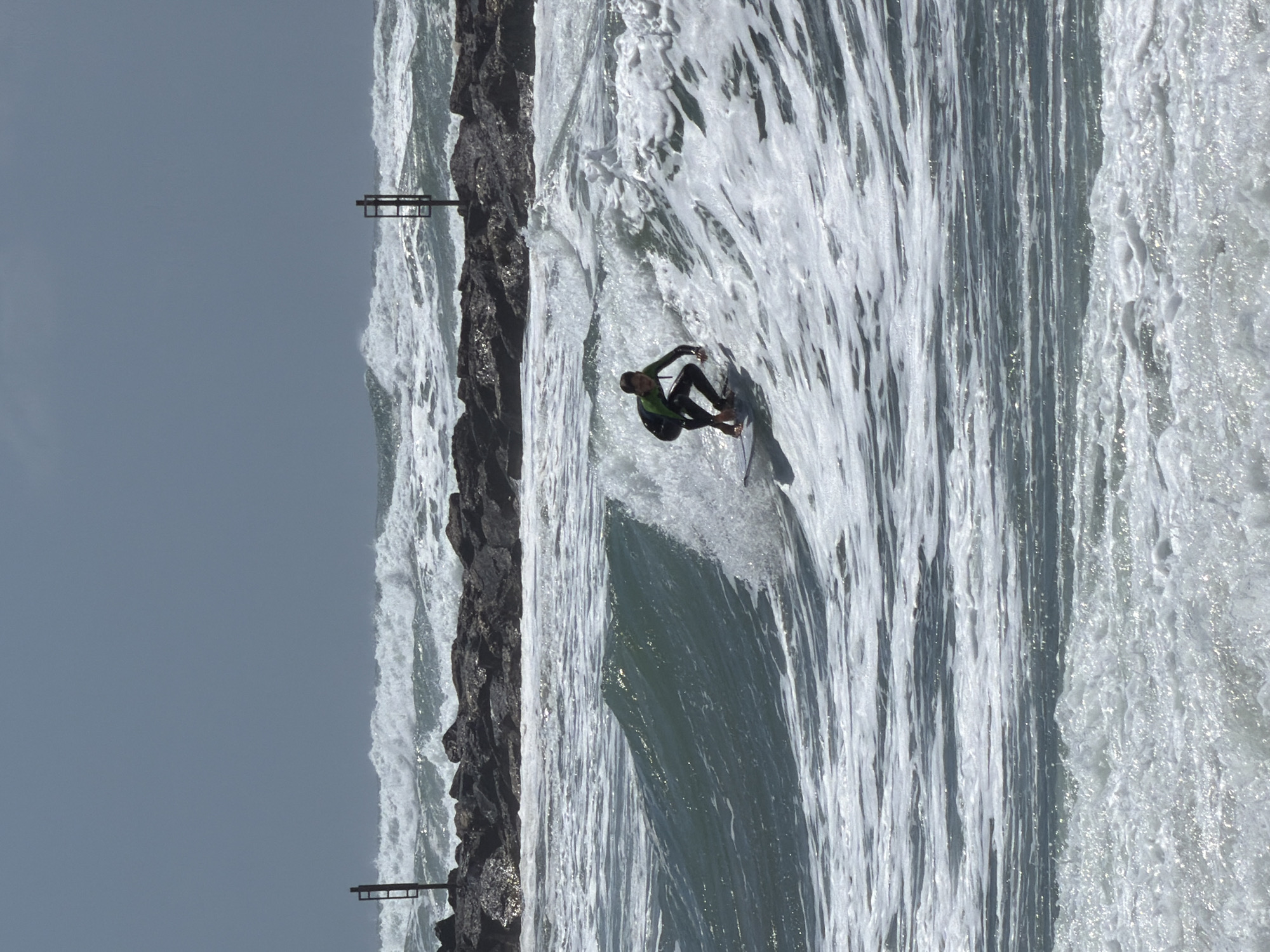 A surfer rides a wave near a breakwater in Tel Aviv, Israel. The breakwater is composed of dark gray rocks and is lined with a simple metal fence. The sea is turbulent with white foam and spray. The surfer, wearing a dark wetsuit with green accents, is positioned centrally on the wave. The sky is overcast and gray.