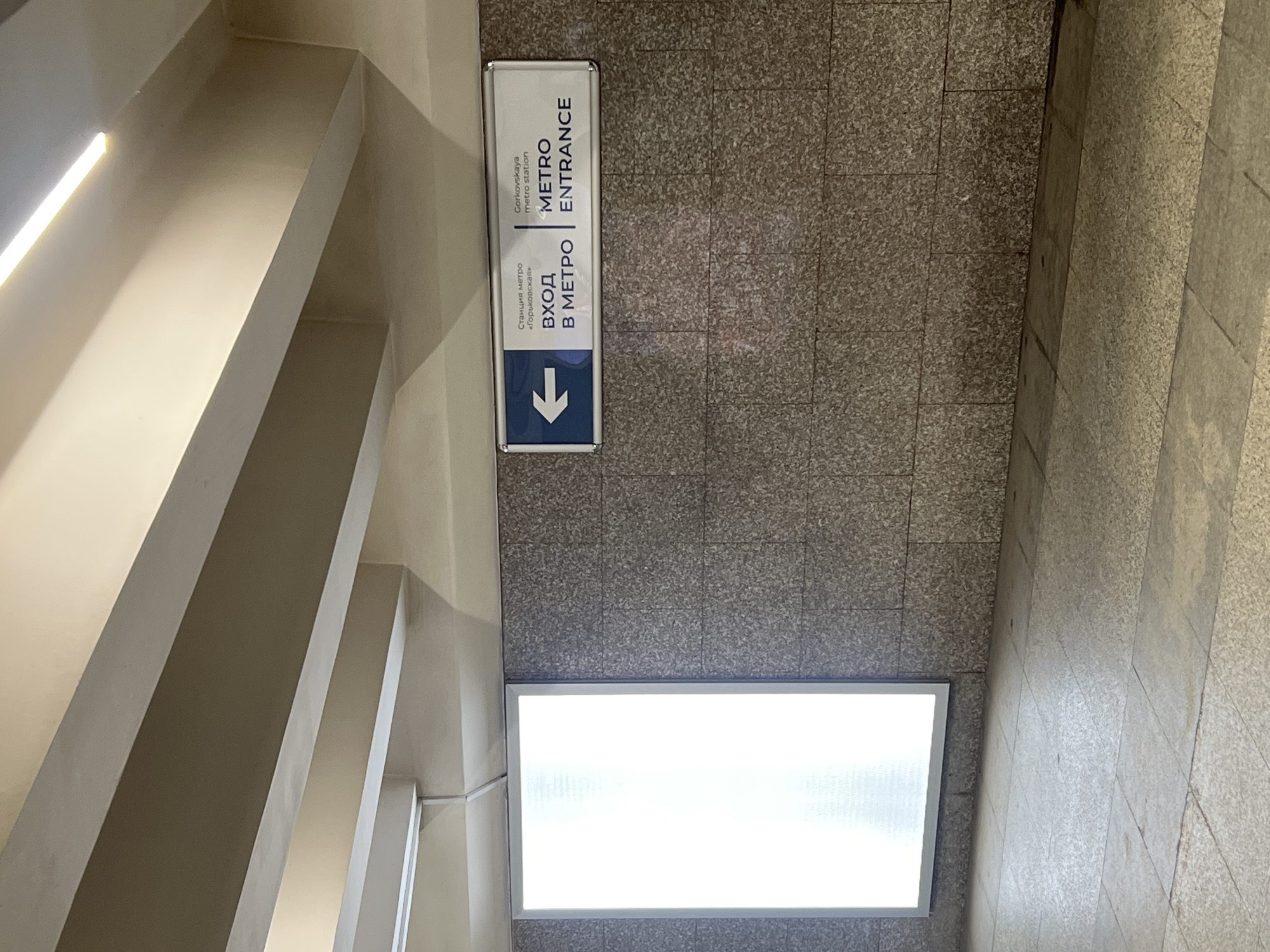 The image shows a subway entrance sign in Nizhny Novgorod, Russia. The sign is blue and white and has an arrow pointing to the left. The text on the sign is in both Russian and English and reads "Gorkovskaya metro station METRO ENTRANCE". The sign is mounted on a textured gray wall. To the left of the sign is a large, brightly lit rectangular panel. The floor is made of large square tiles. The ceiling is made of concrete beams. The overall impression is that of an underground passageway or station.