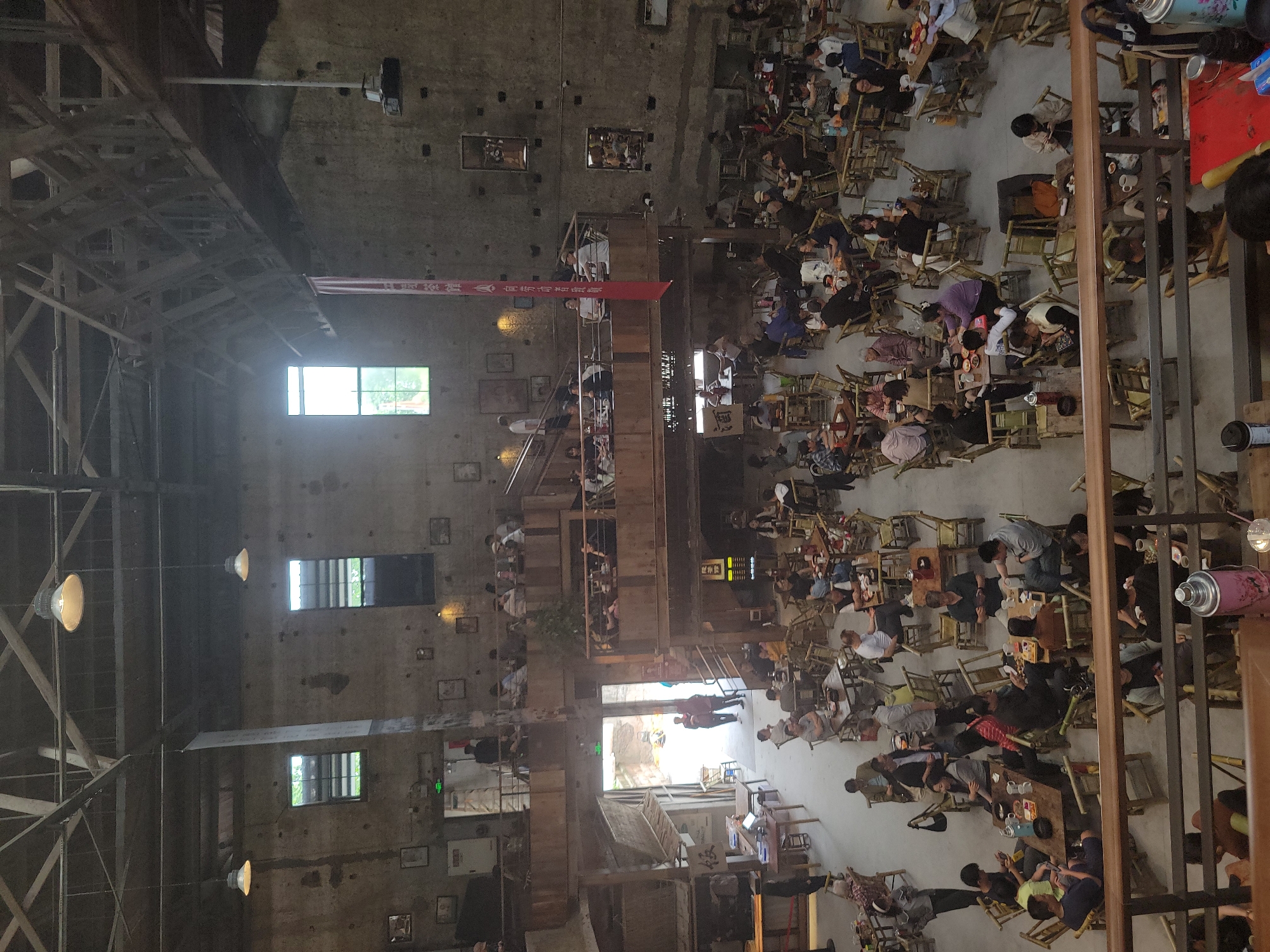The image shows the interior of a large, industrial-style space filled with people seated at numerous small tables. The setting is a multi-level structure with exposed beams, brick or concrete walls, and several windows admitting natural light. On the upper levels, people are seated along balconies or walkways overlooking the main floor. Red banners with Chinese characters are visible hanging within the space. Individuals are engaged in various activities, primarily eating, drinking, and socializing. The seating arrangement is dense, indicating a crowded or busy environment. The image suggests the scene is in the city of Liangjiang, China.