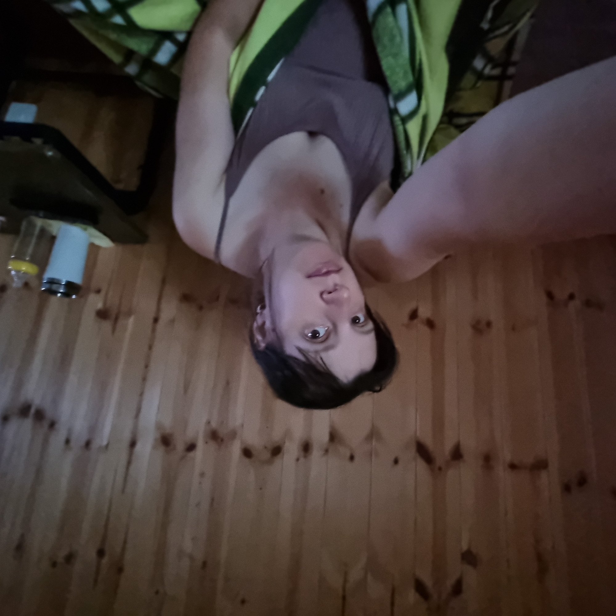 A woman with dark hair and light skin is shown from the chest up, holding a camera to take a selfie. She is wearing a grey spaghetti strap tank top and a green and yellow plaid blanket is wrapped around her. She is sitting in front of a wood-paneled wall. To the right of the woman, on a dark table, are a white insulated tumbler and a baby bottle. The scene appears to be indoors.