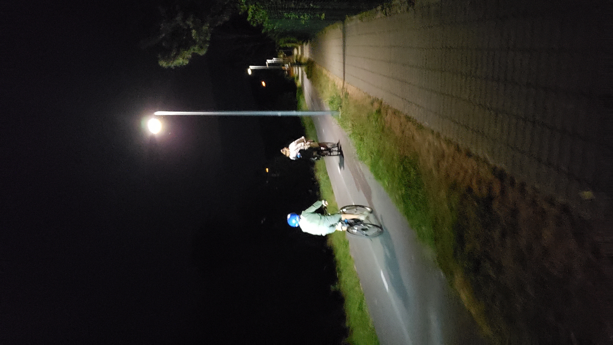 The image depicts a scene at night on a paved pathway and adjacent bicycle lane, illuminated by a streetlamp. Two individuals are riding bicycles. The person in the foreground, on the bicycle lane, is wearing a light-colored jacket and a blue helmet. The second person, further ahead on the bicycle lane, is wearing a light-colored shirt and dark pants. The pathway is to the right of the bicycle lane and is paved with interlocking bricks. Grass and foliage are visible to the left of the bicycle lane, and a fence line runs along the right edge of the pathway. The overall environment is dark, suggesting it is nighttime. The location is not specified in the image or its context, therefore, it cannot be definitively stated to be in Prešov, Slovakia.