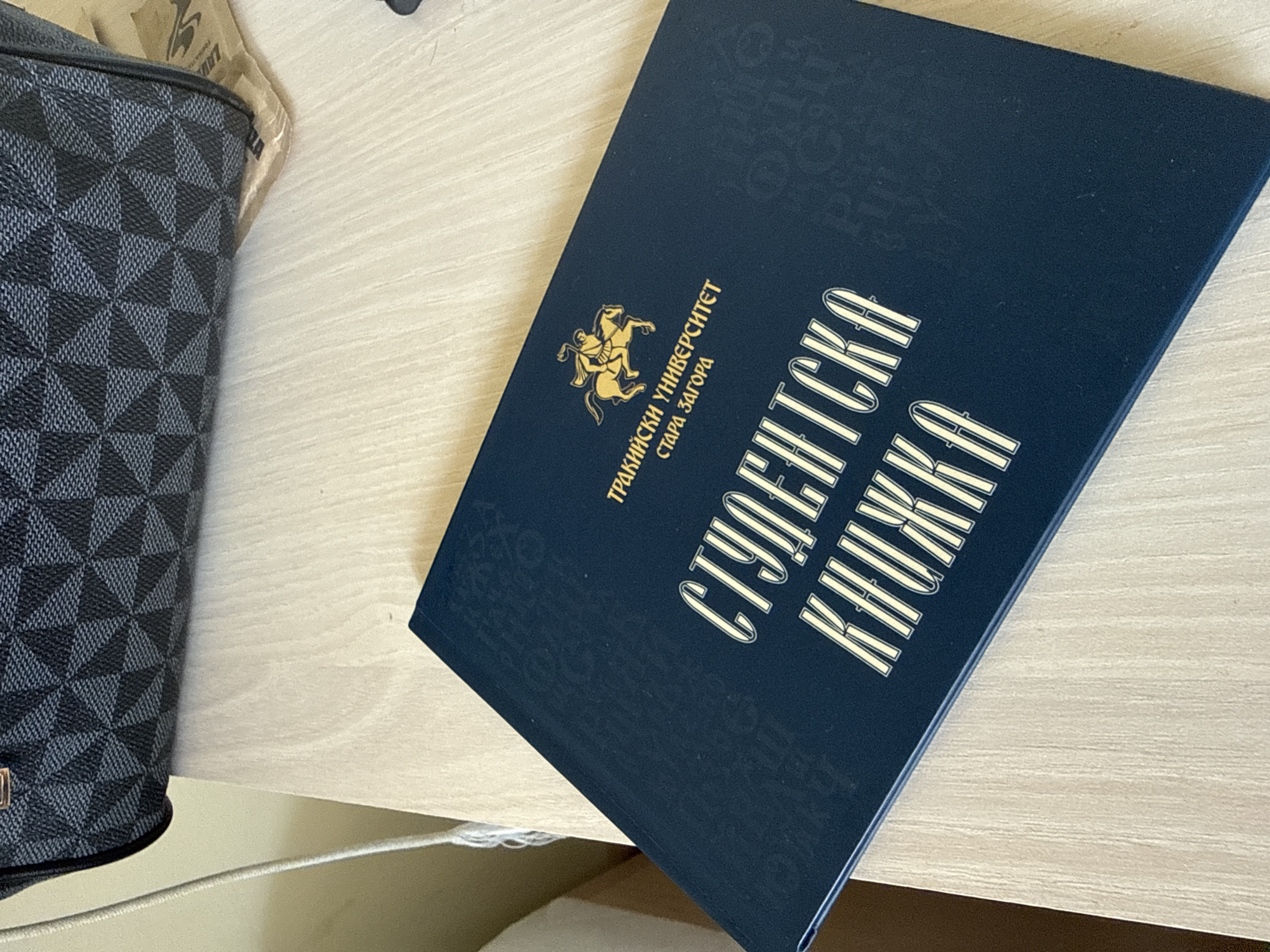 The image displays a close-up, slightly angled view of a dark blue book and a patterned bag resting on a light-colored wooden surface.
The most prominent object is a dark blue book positioned on the right side of the frame. Its cover faces upwards, angled towards the top right. Emblazoned in gold on the cover is an emblem featuring two rearing horses and a rider. Below the emblem, arranged vertically, is the text "ТРАКИЙСКИ УНИВЕРСИТЕТ" (Trakian University) followed by "СТАРА ЗАГОРА" (Stara Zagora). Horizontally across the main part of the cover, in large, stylized gold capital letters, are the words "СТУДЕНТСКА КНИЖКА" (Student's Book/Student ID). This text indicates the book is a student's official document from Trakian University, located in Stara Zagora, Bulgaria.
To the left of the book, a dark grey and black bag is partially visible. It features a distinctive geometric pattern composed of interlocking squares and triangles, creating a textured, three-dimensional checkered appearance. The bag appears to be made of a durable material.
Both items are placed on a light wooden surface, likely a table or desk, which has a subtle grain visible. The lighting is soft and even, suggesting an indoor setting. There are no people visible, and no specific activity is taking place. The scene is a simple still life, showcasing these two personal items.