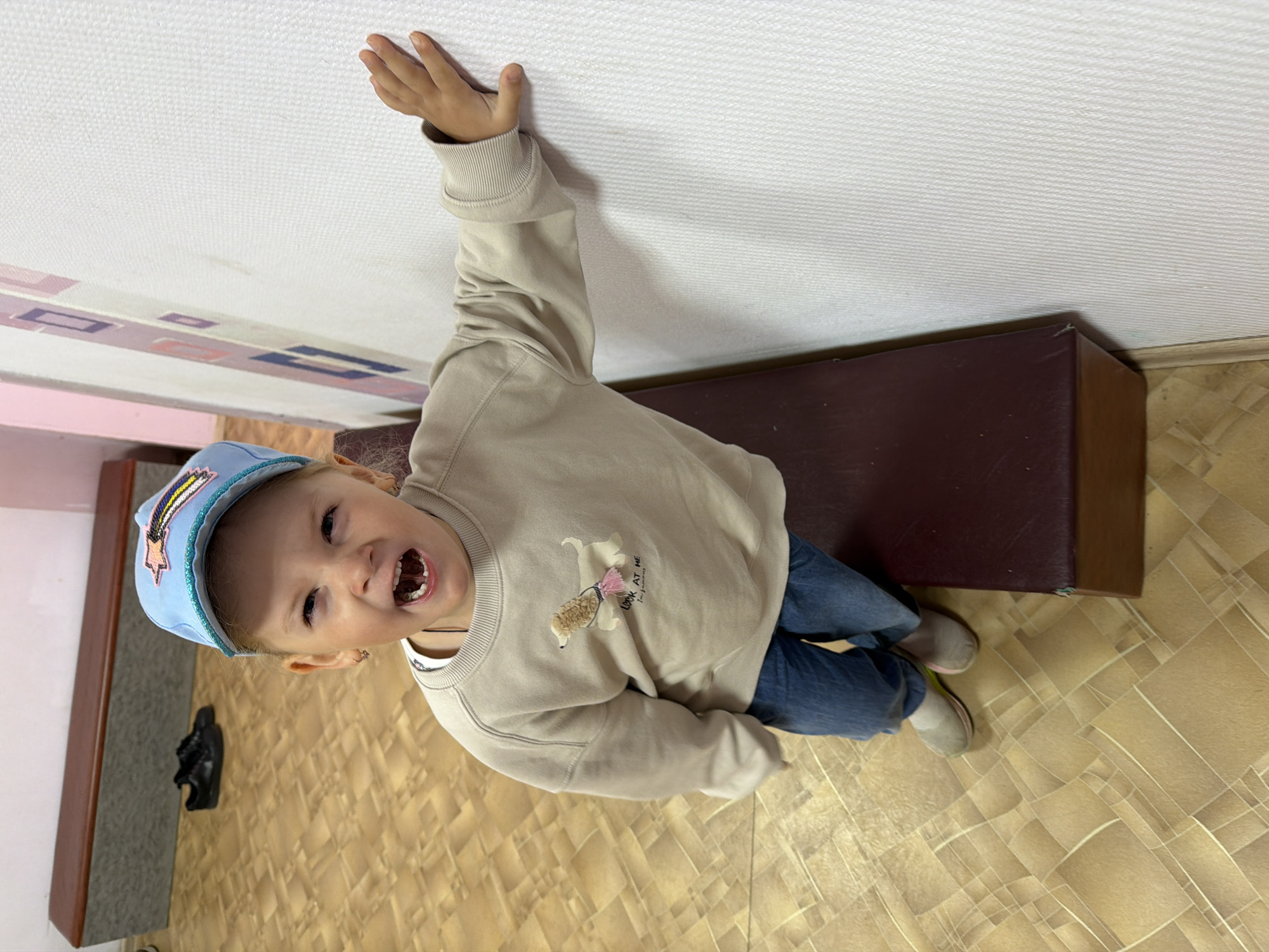 A young girl wearing a blue baseball cap and a light beige sweatshirt with a dog graphic is standing with her right hand on a white textured wall. She is wearing blue jeans and light-colored sneakers. The girl is facing the camera with her mouth open, appearing to be speaking or laughing. She is positioned next to a dark red bench. The floor is tiled with a geometric pattern in shades of brown and yellow. In the background, a pair of black sneakers are visible on the floor near a longer bench. The environment suggests an indoor public space, potentially a waiting area or a hallway.
