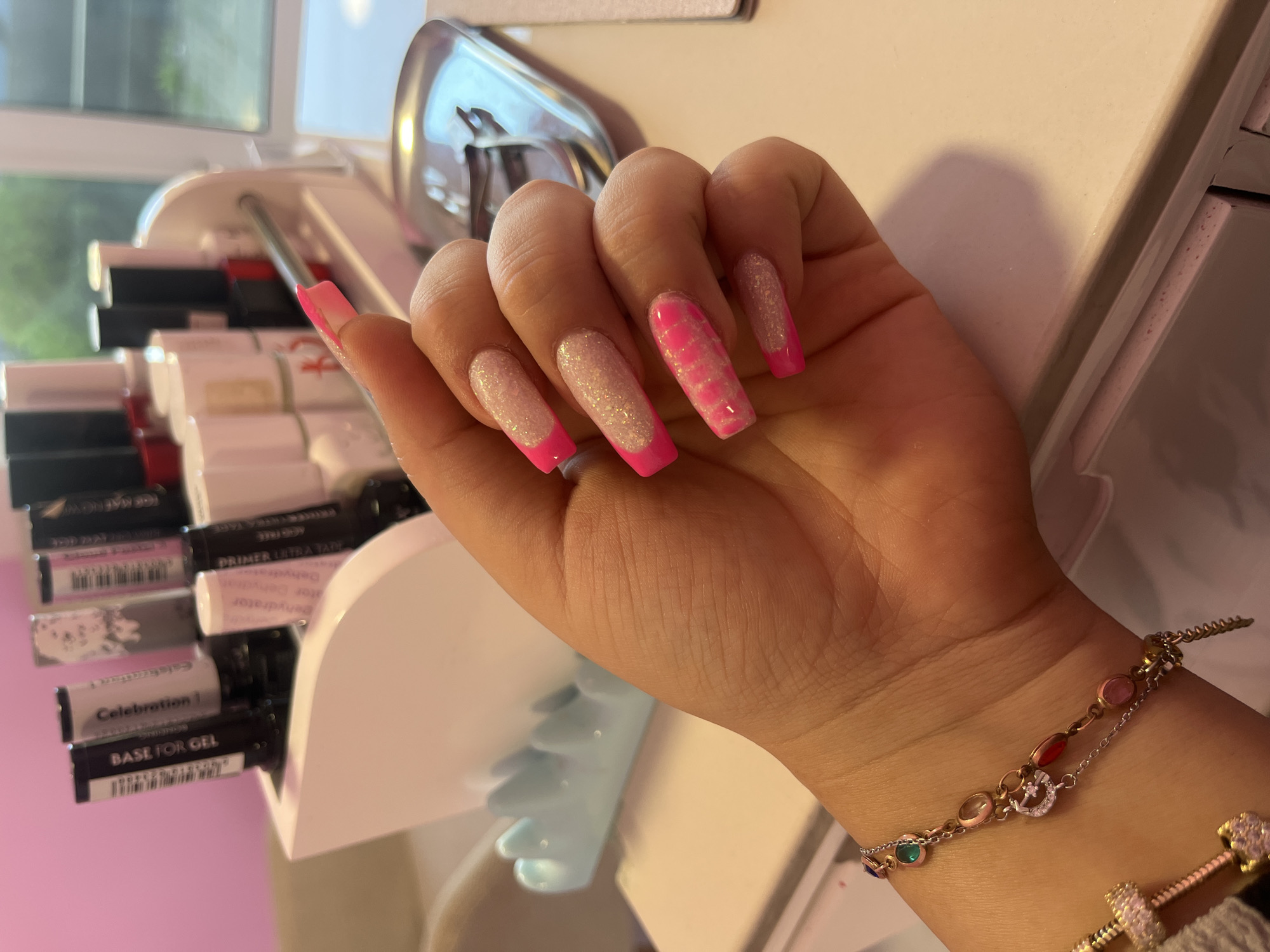 The image shows a close-up of a person's left hand, displaying brightly colored acrylic nails with a French tip design. The nails are long and have a squared-off shape. The nail polish colors include hot pink and white with glitter. The hand is resting on a white surface, possibly a vanity or desk, with a blurred background that appears to be a nail salon. In the background, various nail polish bottles and salon equipment are visible, arranged in a white organizer. A delicate bracelet with colorful charms is worn on the wrist. The overall impression is of a professional nail service being showcased. There is no indication of the city of Novi Pazar, Bulgaria in the image.