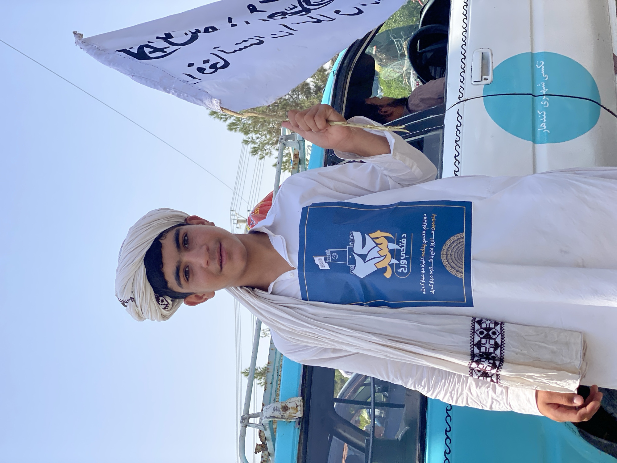 A young boy, likely a teenager, stands in front of a light blue and white car in Kandahar, Afghanistan. He is wearing a white turban and a white tunic with a dark embroidered trim on the sleeves. A dark blue placard with white and yellow lettering and an illustration of a fortified tower and a hand is attached to his tunic. He holds a white flag with black Arabic script in his right hand. The car has a blue circular emblem on the side with white text. The background shows a clear sky, trees, and buildings, suggesting an outdoor urban environment.