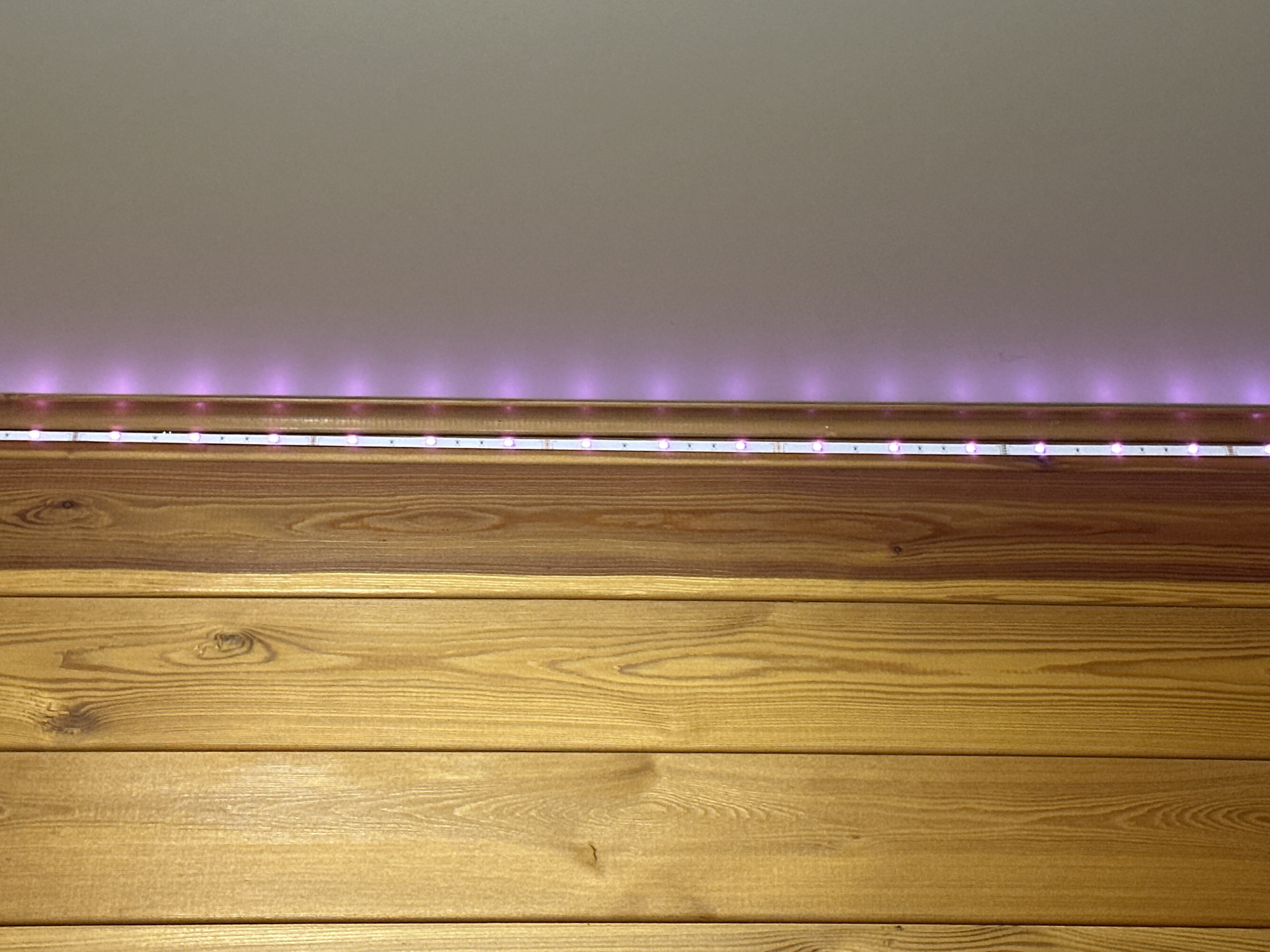 The image shows a close-up view of a wall with horizontally aligned wooden planks. At the top of the wooden planks, a strip of LED lights emits a pinkish-purple glow. The ceiling above the lights is a light gray color. The wood has a warm, reddish-brown hue with visible grain patterns. The lighting appears to be artificial, coming from the LED strip. There are no people or any other objects visible in the frame. The scene is static and appears to be an interior detail of a room.