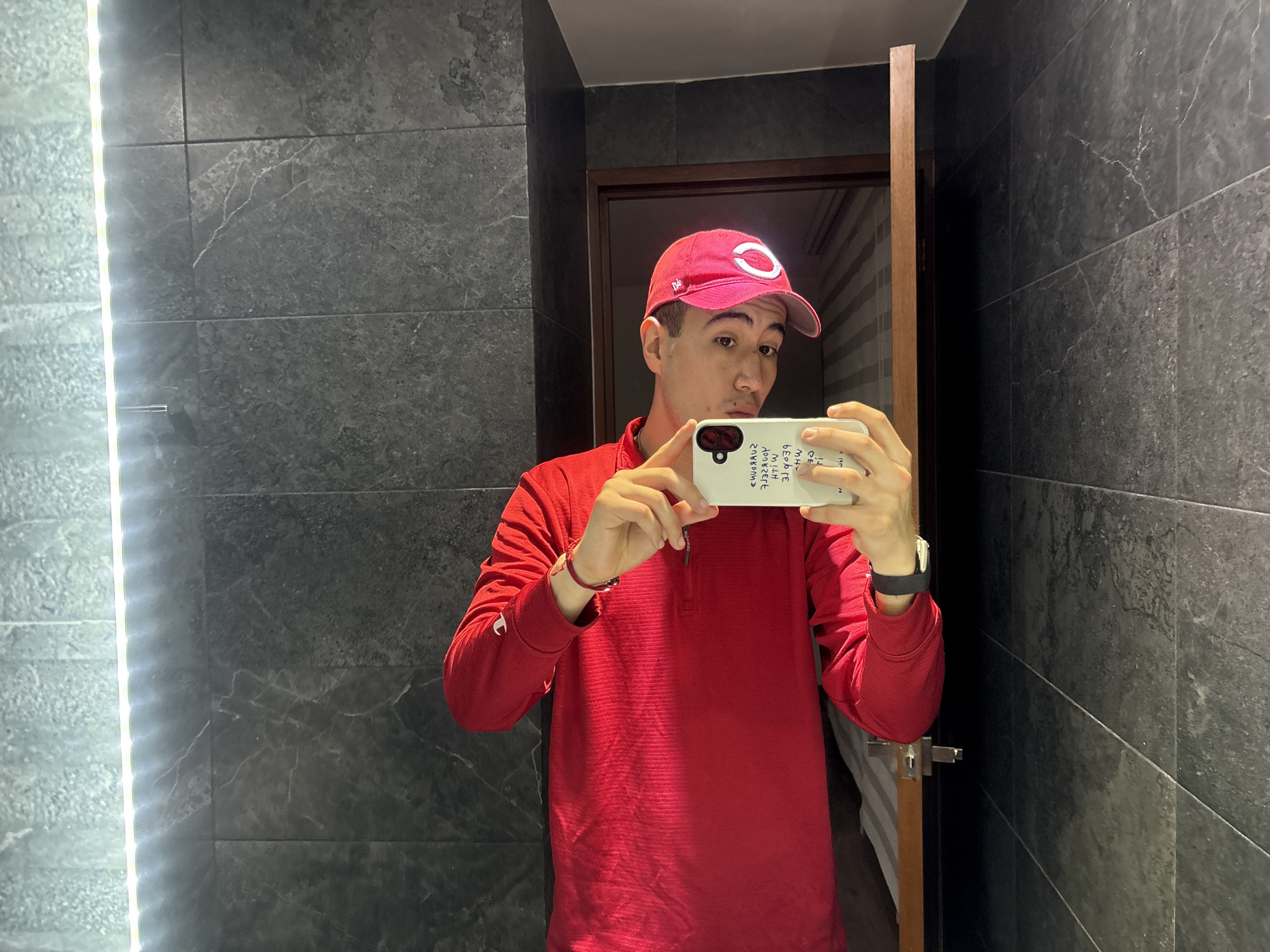 A man is captured taking a mirror selfie indoors, likely in a bathroom, in San Bernardino Tlaxcalancingo, Mexico. He wears a red long-sleeved quarter-zip shirt and a red baseball cap with a prominent white "C" logo. He holds a white smartphone with his right hand, its screen obscured by his hand but showing text and camera details in reverse due to the mirror reflection. A black smartwatch is on his left wrist, and a red string bracelet adorns his right wrist.
The setting features dark grey, possibly marble or granite, tiled walls with subtle white veining. A bright vertical LED light strip runs along the left side of the mirror, illuminating the scene. In the reflection behind the man's right shoulder, a wooden door frame is visible, hinting at another room or hallway beyond. The man has a neutral to slightly pouting expression as he focuses on capturing the image.
Visible text on the back of the white phone case, appearing mirrored, includes phrases and words such as "SWIMMING," "SWIM," "WITH ME," "HI," "GE," "CE," and the number "3046." Additionally, standard camera lens labels like "ULTRA WIDE," "2X TELE," and "1X WIDE" are faintly discernible around the phone's camera module. The lighting is bright and artificial, suggesting an indoor setting.