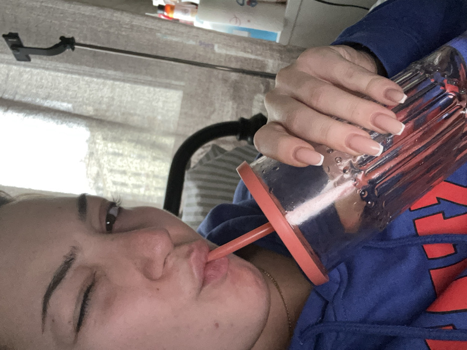 A young female is depicted indoors in Schiltigheim, France, from a frontal, chest-up perspective. She wears a blue hooded sweatshirt with red lettering and a thin gold-colored necklace. Her left eye is closed in a wink, while her right eye remains open and directed towards the viewer. She is actively drinking from a transparent, ribbed plastic tumbler with an orange-red lid and matching straw, holding it with her right hand. The tumbler contains liquid and displays exterior condensation. Her fingernails are manicured with a French tip style. The background includes a bright light source, consistent with a window covered by light-colored sheer curtains. A dark metal bed frame or chair back is partially visible to her left. To the right, a black extendable pole, resembling a selfie stick or stand, is present, alongside a partial view of a white storage unit holding various bottles.