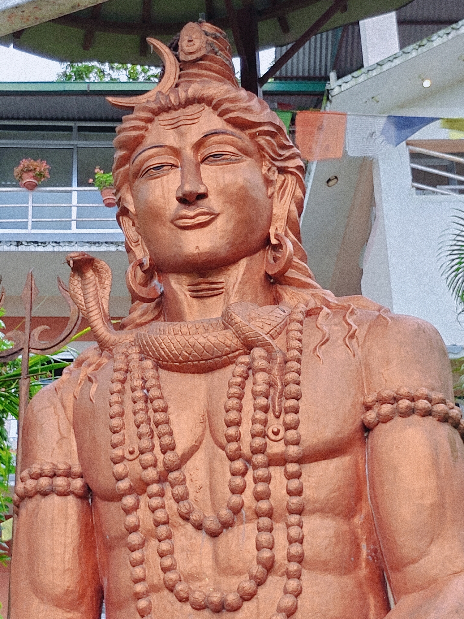 A close-up, eye-level, medium shot captures a large copper-colored statue of the Hindu deity Shiva. The statue depicts Shiva from the chest up, with the deity's head slightly tilted and looking forward with a serene expression. A crescent moon adorns Shiva's hair, which cascades down the shoulders in sculpted waves. A cobra is wrapped around Shiva's neck and shoulder, with its head raised and poised. Multiple strands of beaded necklaces hang from Shiva's neck, and a beaded bracelet encircles the right wrist. The background features a building with windows and balconies, some with potted plants. Prayer flags, some orange and white, hang from the building. The statue is outdoors, suggesting it is located in a public space or temple complex. While the exact location in Pokhara, Nepal, is not specified, the presence of prayer flags and the style of the building are consistent with religious or cultural sites in the region. The lighting is natural daylight, casting soft shadows on the statue's features.