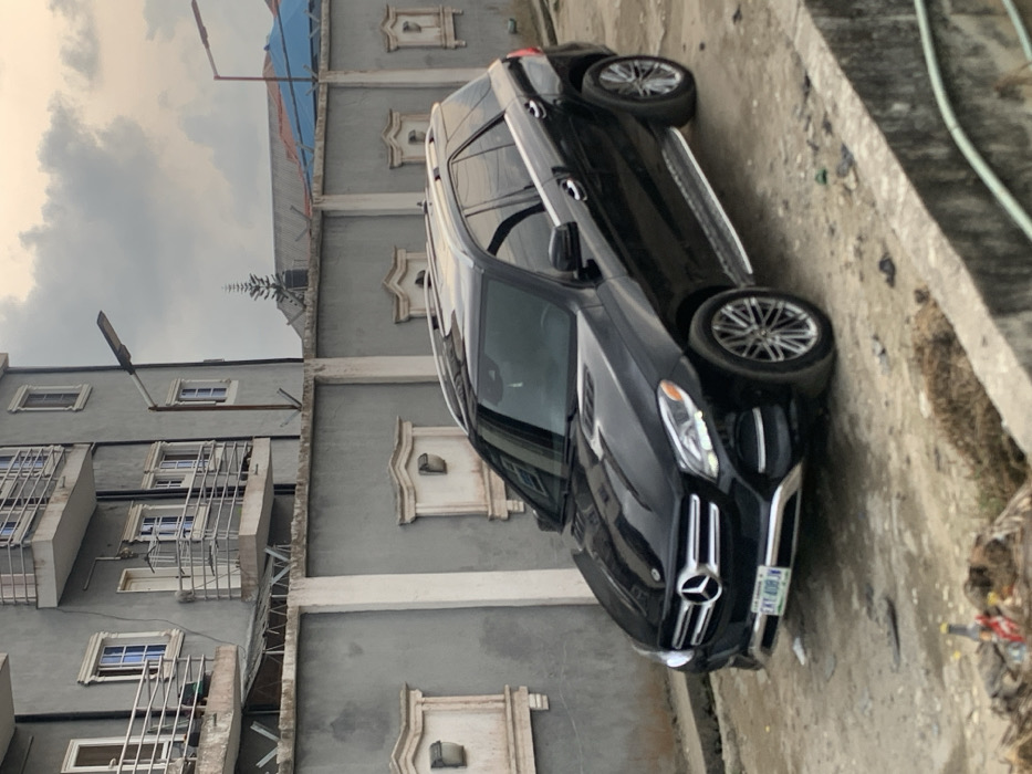 A black Mercedes-Benz GLE SUV, bearing Nigerian license plate "LKL-408JN" (Lekki, Lagos), is parked on a dirt and gravel surface next to a concrete drainage ditch in Lekki, Nigeria. The vehicle faces slightly left, its front bumper and grille visible, reflecting the overcast sky. A multi-story residential building, painted grey with multiple white-framed windows and balconies featuring metal railings, stands behind a tall, grey concrete perimeter wall topped with razor wire. The wall incorporates decorative rectangular recesses, each containing a solar-powered wall light. Two streetlights with LED fixtures are visible: one attached to the residential building's wall and another further in the background. A conical evergreen tree and a blue tarp-covered roof are visible over the perimeter wall. No individuals are present in the scene. Debris, including a plastic bottle, is scattered near the drainage ditch in the foreground. The sky is largely overcast with grey clouds.