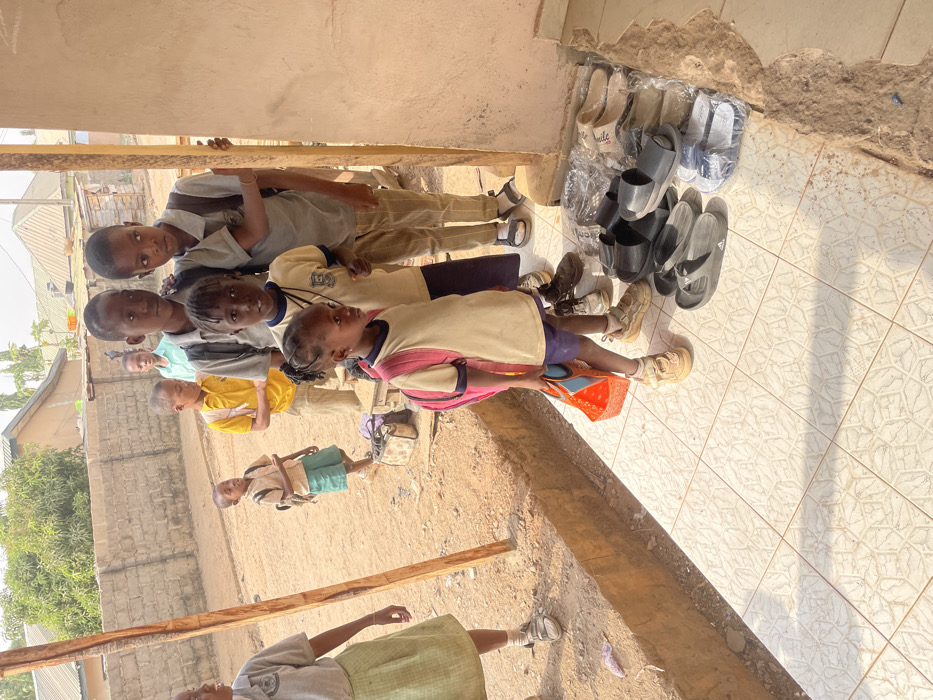 The image shows a group of school-aged children gathered outdoors, appearing to be in a line or congregation outside a building. The image is oriented vertically, with the ground surface appearing primarily on the right side and stretching towards the bottom, while the children and buildings occupy the left and central parts of the frame.
In the immediate foreground, on the lower right, there is a patterned tiled floor where several pairs of shoes, sandals, and slippers are neatly arranged against a light-colored wall. Many of these are black, while some are lighter in color, and a few are still wrapped in clear plastic. This arrangement suggests footwear might be removed before entering the adjacent building.
To the left of the tiled area, the ground transitions to a dirt or sandy surface. On this surface, a group of children is standing.
The closest child in this group, positioned centrally, is a young girl wearing a cream-colored top with pink sleeves and blue shorts, holding a small orange bucket or container. She is looking towards her left. Her feet are in light brown shoes.
Beside her, slightly to her left, is another young girl with braided hair, wearing a white collared shirt (possibly a school uniform with a visible logo on the left chest) and dark trousers. She is looking directly at the camera.
Behind these two, several other children are visible. One boy wears a similar white collared shirt and dark trousers, also looking towards the camera. Further back and slightly to the left, another boy in a grey collared shirt and light trousers is also looking at the camera. A young boy in a yellow short-sleeved shirt stands further back in the group. In the far background, a child in a light blue shirt is partially visible.
At the very bottom left of the frame, a person (likely an adult) is partially visible, wearing a light green skirt and a white top.
The background consists of concrete block walls of buildings and some distant sparse vegetation, suggesting a semi-urban or rural setting. A rough wooden beam or pole is lying horizontally on the dirt ground in the lower-left portion of the image.
The scene is brightly lit by strong sunlight, casting distinct shadows on both the tiled floor and the dirt ground, indicating a clear, sunny day, likely morning or late afternoon given the angle of the shadows. The children generally have neutral or slightly curious expressions. No text is visible in the image.