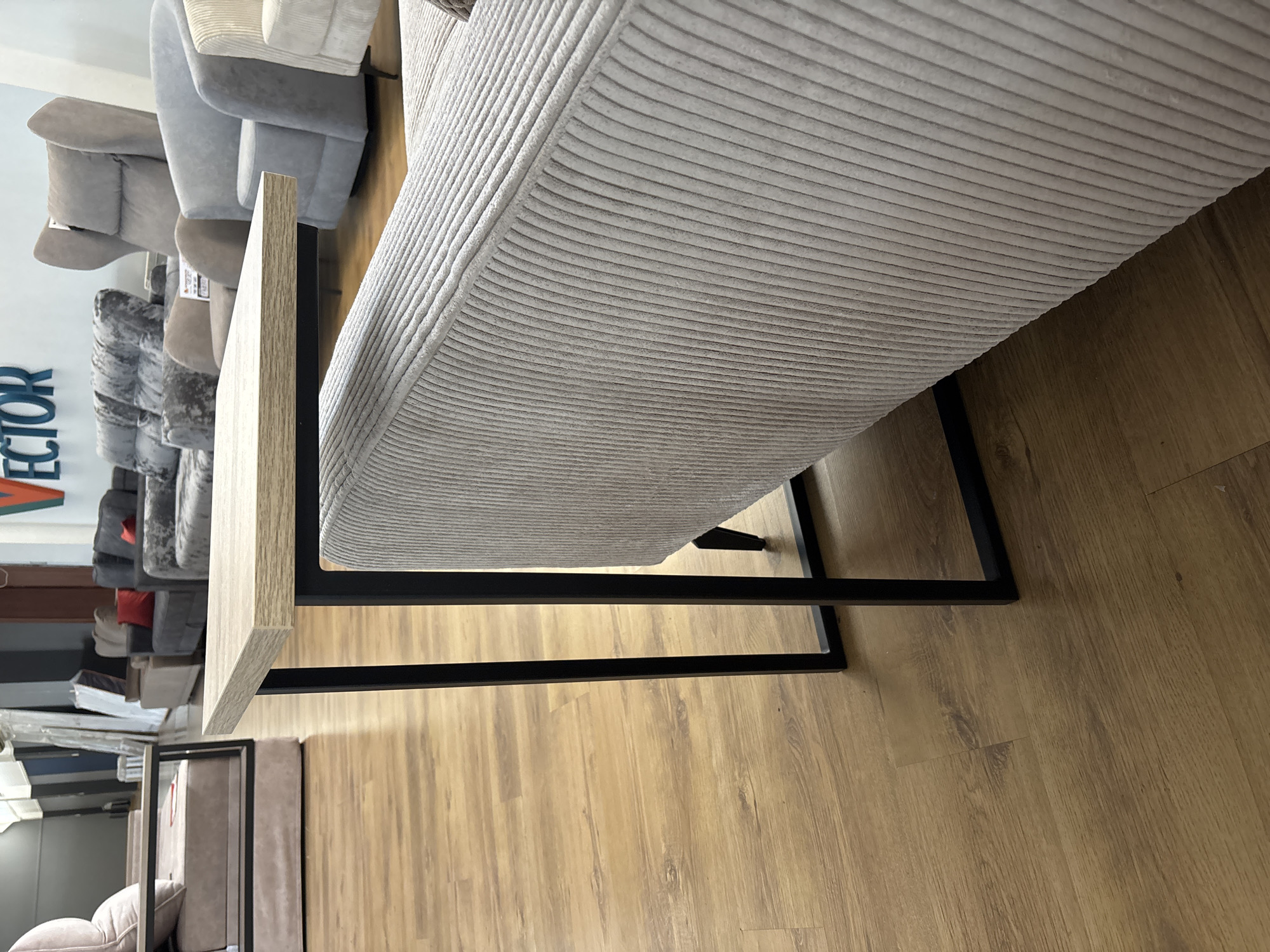 The image depicts a furniture store in Zaporizhzhia, Ukraine, showcasing a variety of sofas and armchairs. In the foreground, a light-colored wooden table with black metal legs is positioned next to a light gray corduroy sofa. The table's surface is angled towards the viewer, highlighting its texture. Behind the sofa, other seating arrangements are visible, including a large sectional sofa with plush cushions and red throw pillows, and several individual armchairs in various shades of gray and beige. The flooring is a light-colored wood laminate. The background reveals a store environment with display racks and what appear to be wrapped furniture items. A sign with the word "VECTOR" in blue lettering is mounted on the wall. There are no people visible in the image.