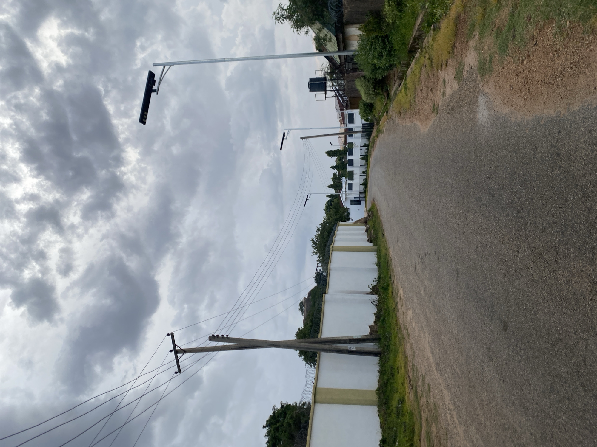 A paved road curves gently to the right in an urban environment under a cloudy sky. On the left side of the road, a white wall topped with barbed wire runs along the property line. A wooden utility pole with multiple wires extends from behind the wall. Further down the road on the left, several modern white buildings with dark-trimmed windows are visible, one with a black-roofed structure. On the right side of the road, a tall, slender metal pole with a solar-powered street light is positioned. Beyond it, another similar street light is seen further down the road. A dark water tank on a metal stand is visible behind a fence on the right, adjacent to a building with a green roof. Vegetation, including trees and bushes, lines the sides of the road and the properties. The overall impression is of a well-maintained residential or commercial area in Barikin Jirga Summa, Nigeria.