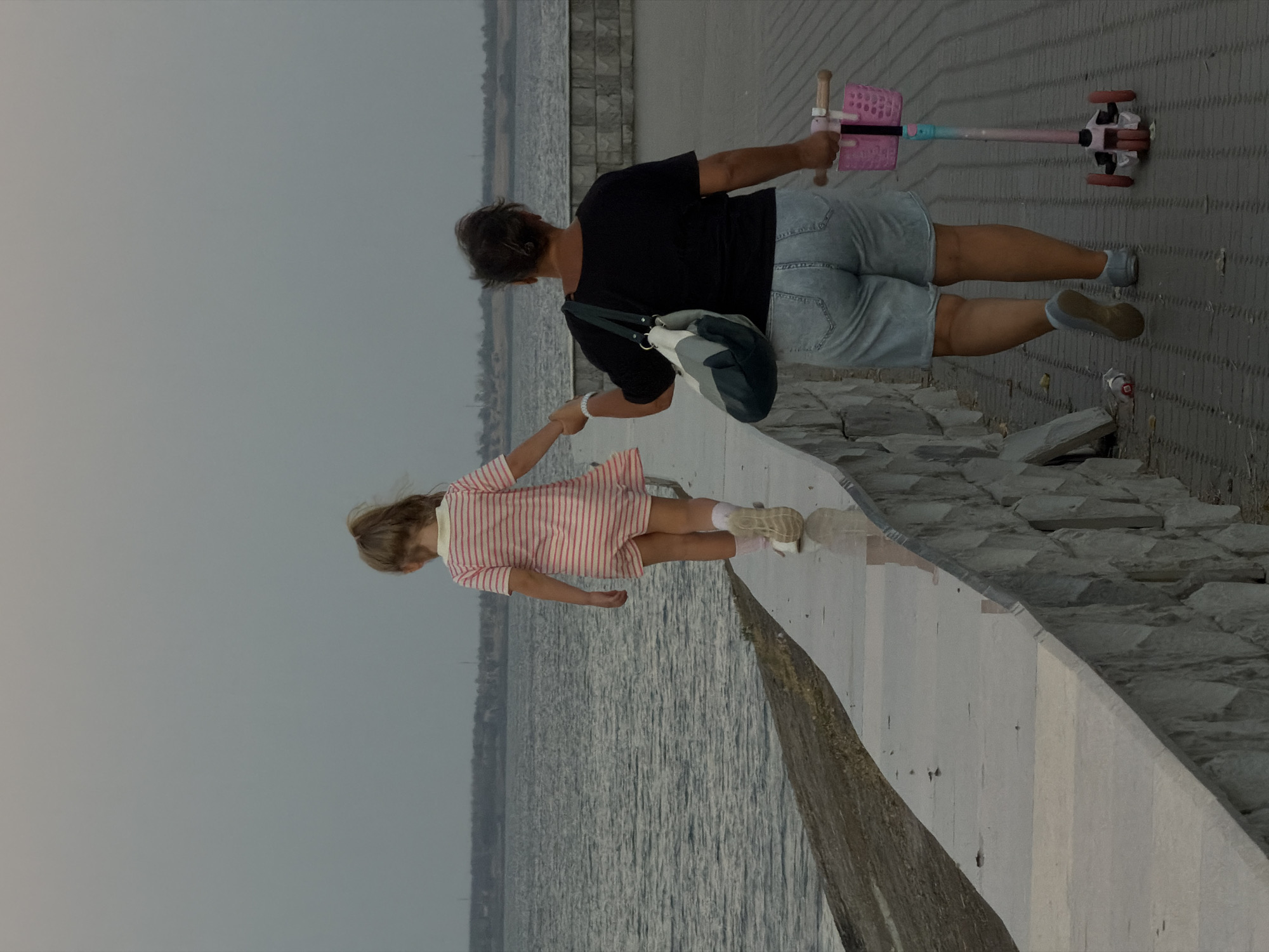 In Kamyshin, Russia, on an overcast day, a woman and a young girl walk along a paved embankment beside a body of water. The girl, facing away from the camera and wearing a pink and white striped dress, walks along the water's edge. The woman, also with her back to the camera, is dressed in a black t-shirt and light blue denim shorts. She carries a multi-colored shoulder bag and walks on the paved walkway behind the girl, holding the handlebars of a pink and blue three-wheeled scooter. The embankment is bordered by a stone wall on the right and a sloped embankment covered in dirt and grass on the left. The sky is uniformly grey and the water appears calm.