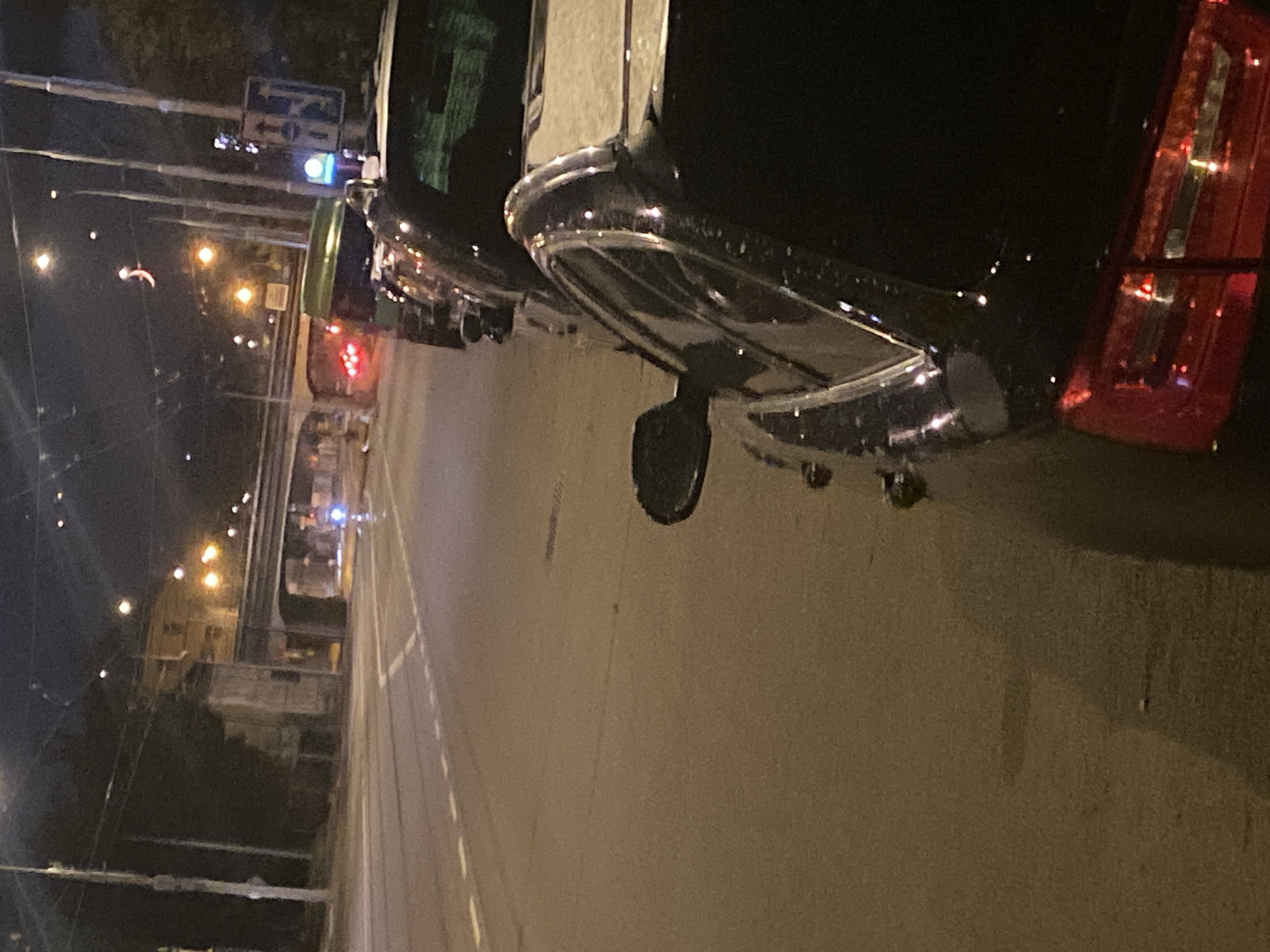 The image depicts a dimly lit urban street scene at night, likely in Brno, Czechia, given the presence of Czech traffic signs. The foreground is dominated by the rear of a dark-colored car, partially illuminated by streetlights and the glow of its red taillights. To the left and extending into the distance, a road with lane markings is visible. In the mid-ground, several other vehicles are parked along the right side of the road. Beyond the parked cars, an elevated structure, possibly a bridge or overpass, spans across the street. The background shows buildings and trees, with streetlights and traffic lights dotting the scene. The overall atmosphere is nocturnal and somewhat hazy due to the low light conditions.
