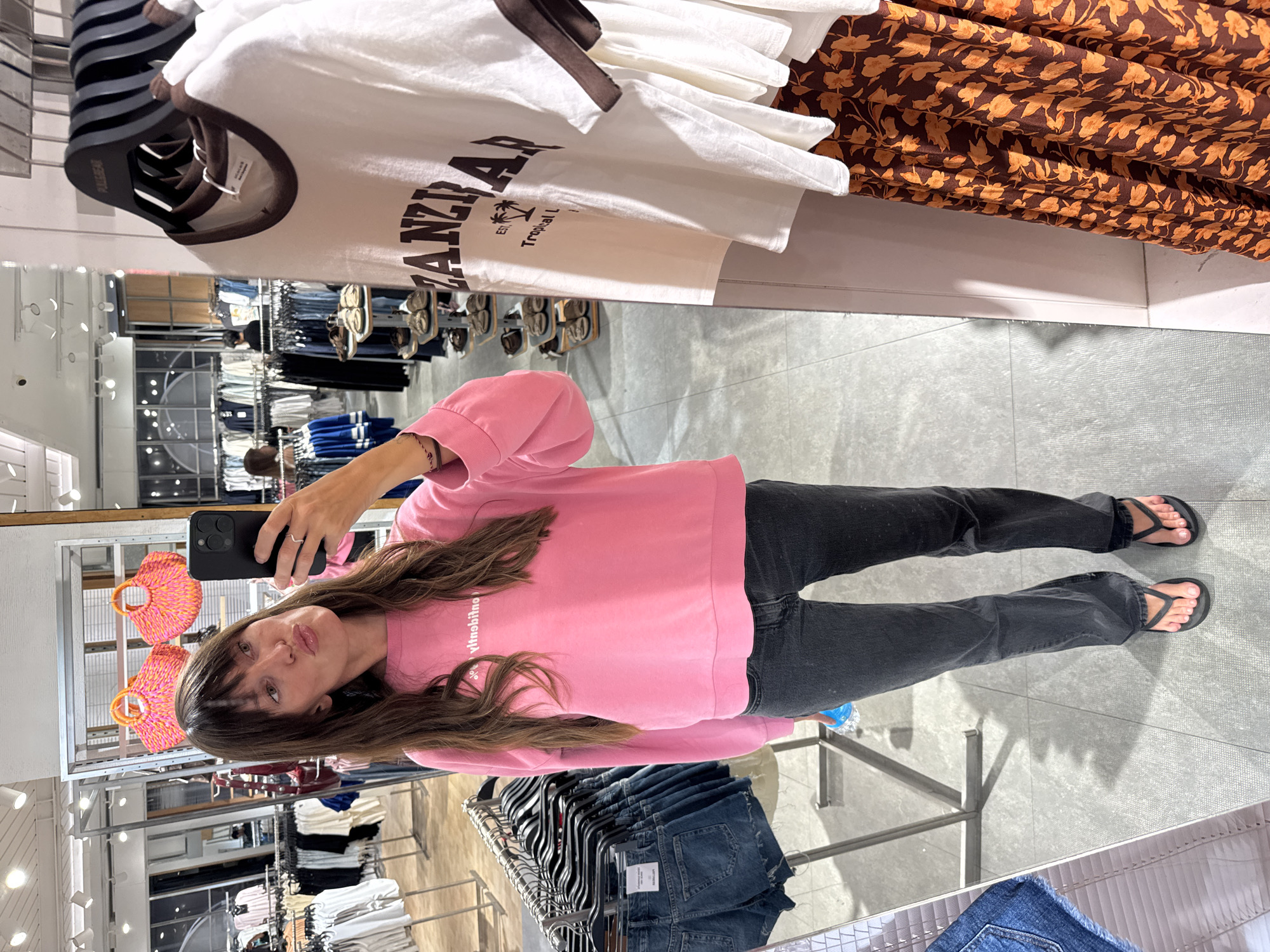 A woman is taking a mirror selfie inside a store. She is wearing a pink sweatshirt, dark gray jeans, and flip-flops. She is holding a black smartphone in her right hand. The store appears to be a clothing store with racks of clothes visible in the background. T-shirts and dresses are on display on racks. The setting resembles a retail environment. The city of Kuta, Indonesia, is not evident in the image.