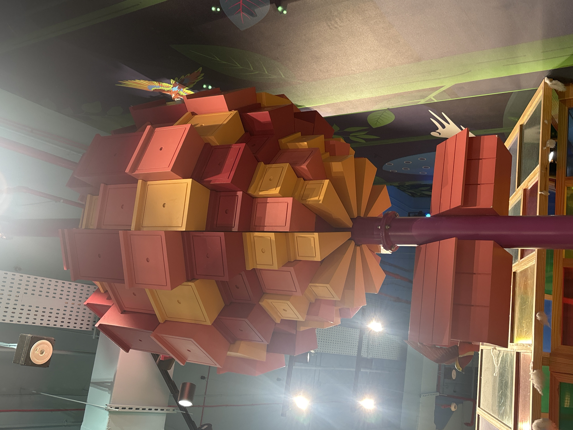The image depicts an interior view of an exhibit likely designed for children, possibly at a museum or a family entertainment center. The central focus is a large, spherical structure composed of numerous colorful, box-like compartments in red and yellow, arranged in a spiraling pattern around a central purple pole. These compartments resemble birdhouses or small rooms.
Below this main structure, red, house-shaped units are attached to the purple pole, one of which has a decorative bird figurine with yellow and red plumage perched on its roof. These units sit above a wooden structure with multiple glass panes, some of which are colored with a rainbow effect, suggesting a display or interactive element.
The background features a dark wall with stylized green foliage and a colorful, flying parrot illustration. The ceiling above shows industrial lighting fixtures and exposed infrastructure, including perforated metal panels and wiring. The overall impression is a vibrant, playful, and educational environment.
This exhibit is located at the Children's City in Dubai, United Arab Emirates.