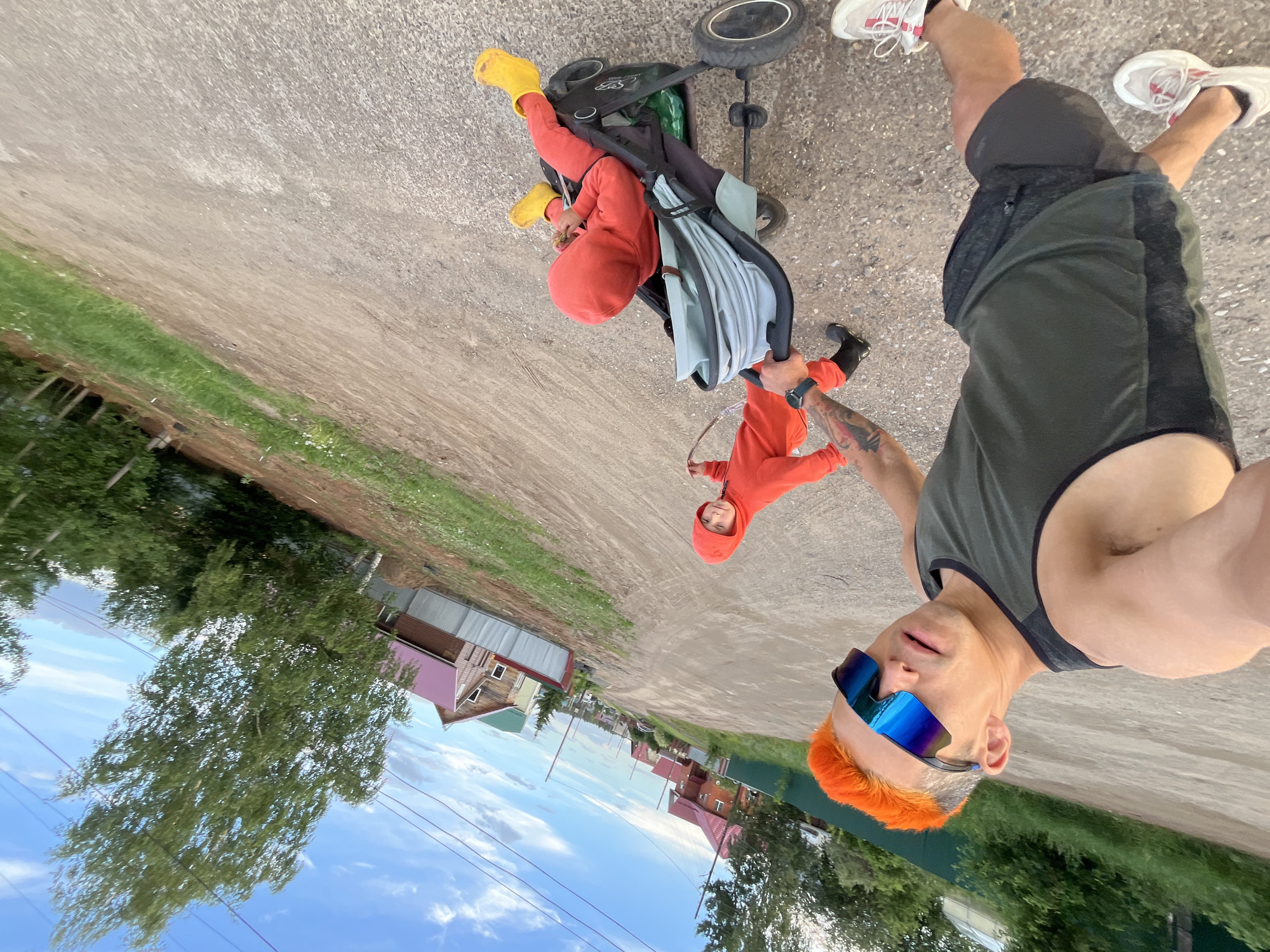 The image is taken at ground level on a road in Chaikovsky, Russia, showing a man taking a selfie. He has orange hair, wears blue sunglasses, a dark green tank top, and dark shorts with white running shoes. He is holding a black stroller with a child dressed in a red hooded outfit and yellow boots inside. Another child, similarly dressed in a red hooded outfit and black boots, is holding the stroller’s handle. The road is gray, and houses are visible in the background under a blue and cloudy sky. Trees are present on either side of the road.