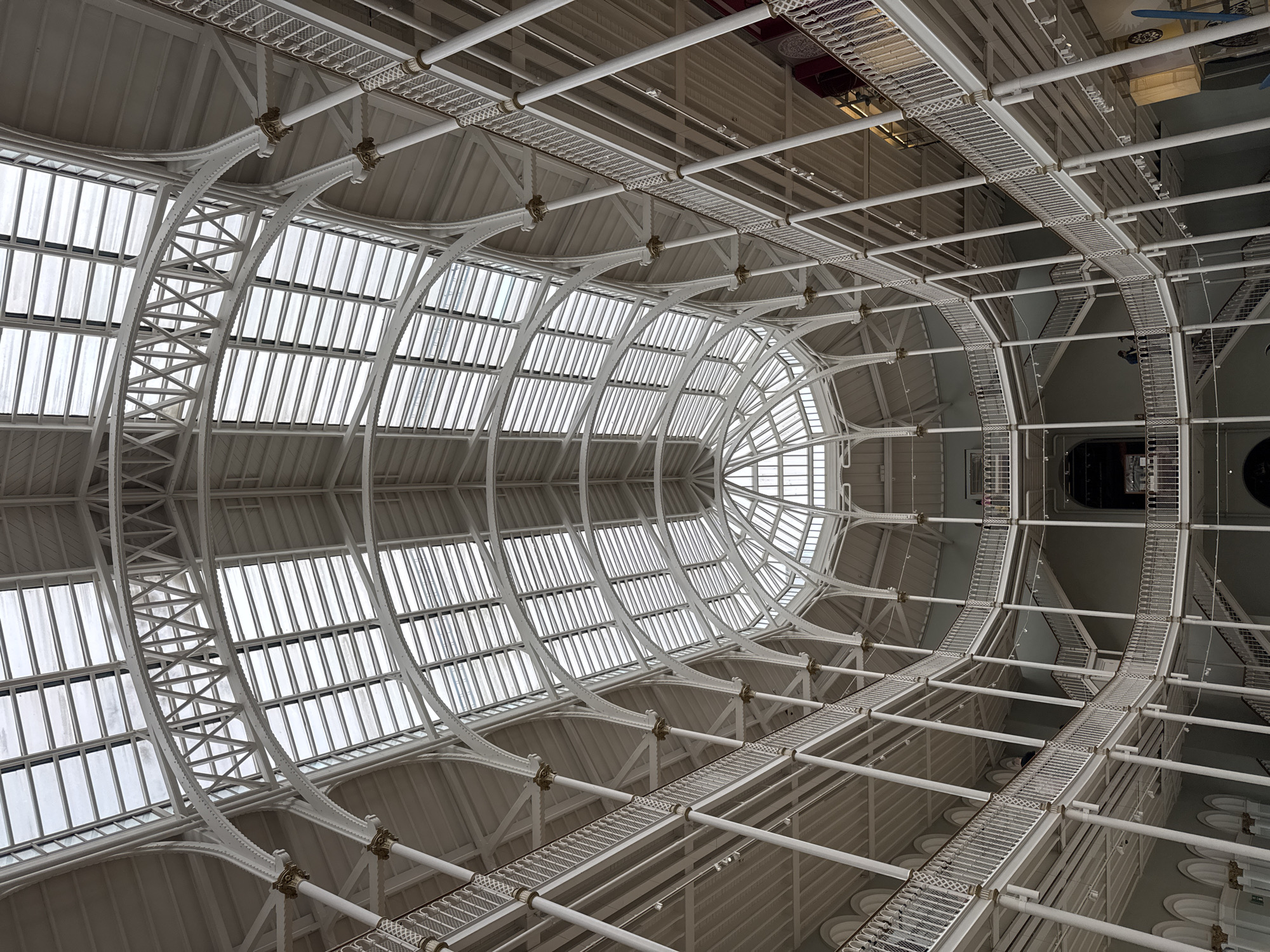 The image shows the interior of the National Museum of Scotland in Edinburgh, United Kingdom, specifically the Grand Gallery. The architecture is characterized by a vast, multi-level atrium with a distinctive glass and iron roof structure that floods the space with natural light. White painted cast iron columns support multiple curved balconies and walkways that encircle the central space, creating a sense of depth and grandeur. Stairs connect the different levels. There are a few individuals visible on the balconies, appearing to be museum visitors. The overall impression is one of an expansive, well-lit, and architecturally significant interior space.