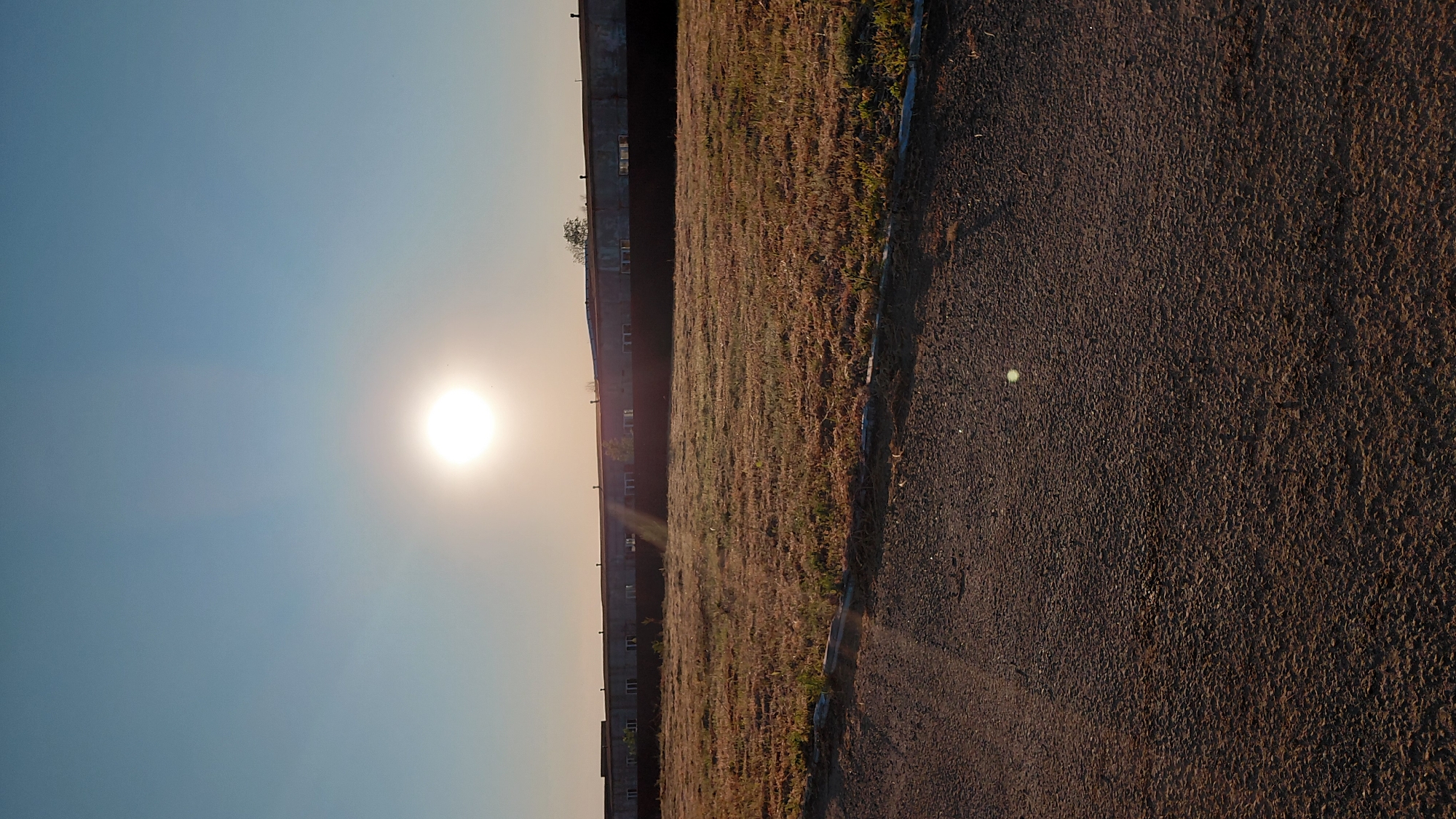The image depicts a rural or semi-urban landscape under a clear, pale blue sky with the sun prominently displayed. The sun is a bright, white disc positioned in the upper left quadrant of the frame, emitting strong rays.
In the middle ground, there is a long, low-lying building with a dark fence in front of it. The building appears to be industrial or a large storage facility, with windows visible along its facade. The fence is dark and solid. Beyond the fence and in front of the building is a patch of dry, yellowed grass.
The foreground consists of a dark, granular surface, likely asphalt or a similar material, with some lighter-colored stones or debris. There is a slight incline, with the dark surface dominating the lower portion of the image. A thin strip of lighter material, possibly a curb or border, separates the dark foreground from the grassy area. A faint lens flare is visible in the lower right quadrant of the image.
The scene appears to be devoid of human presence. The overall atmosphere is one of a sunny day in a somewhat undeveloped or industrial area. The exact location in Saratov, Russia, is not ascertainable from the visual elements of the image alone.