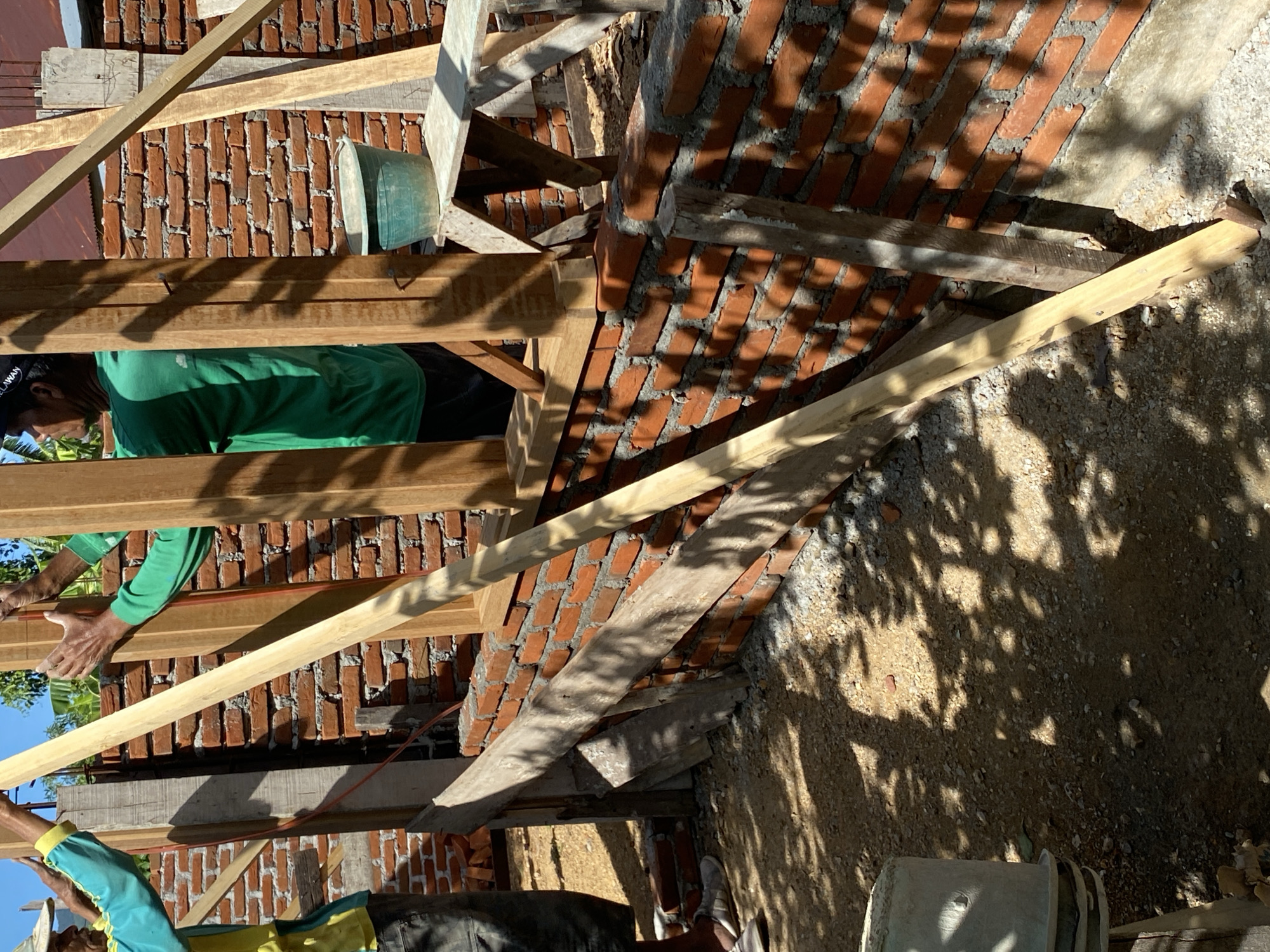 Two construction workers are in the process of building a brick wall. The worker on the left is wearing a yellow and teal long-sleeved shirt and dark pants, while the worker on the right is wearing a green long-sleeved shirt and dark pants, and a black baseball cap. They are standing next to a partially constructed brick wall, with wooden planks and framing visible. One worker is holding a wooden plank upright, while the other is positioned to assist with its placement. In the background, a green bucket sits on a wooden platform next to a brick wall. The ground is covered in gravel and dirt, with shadows cast by nearby foliage. The scene appears to be an outdoor construction site.