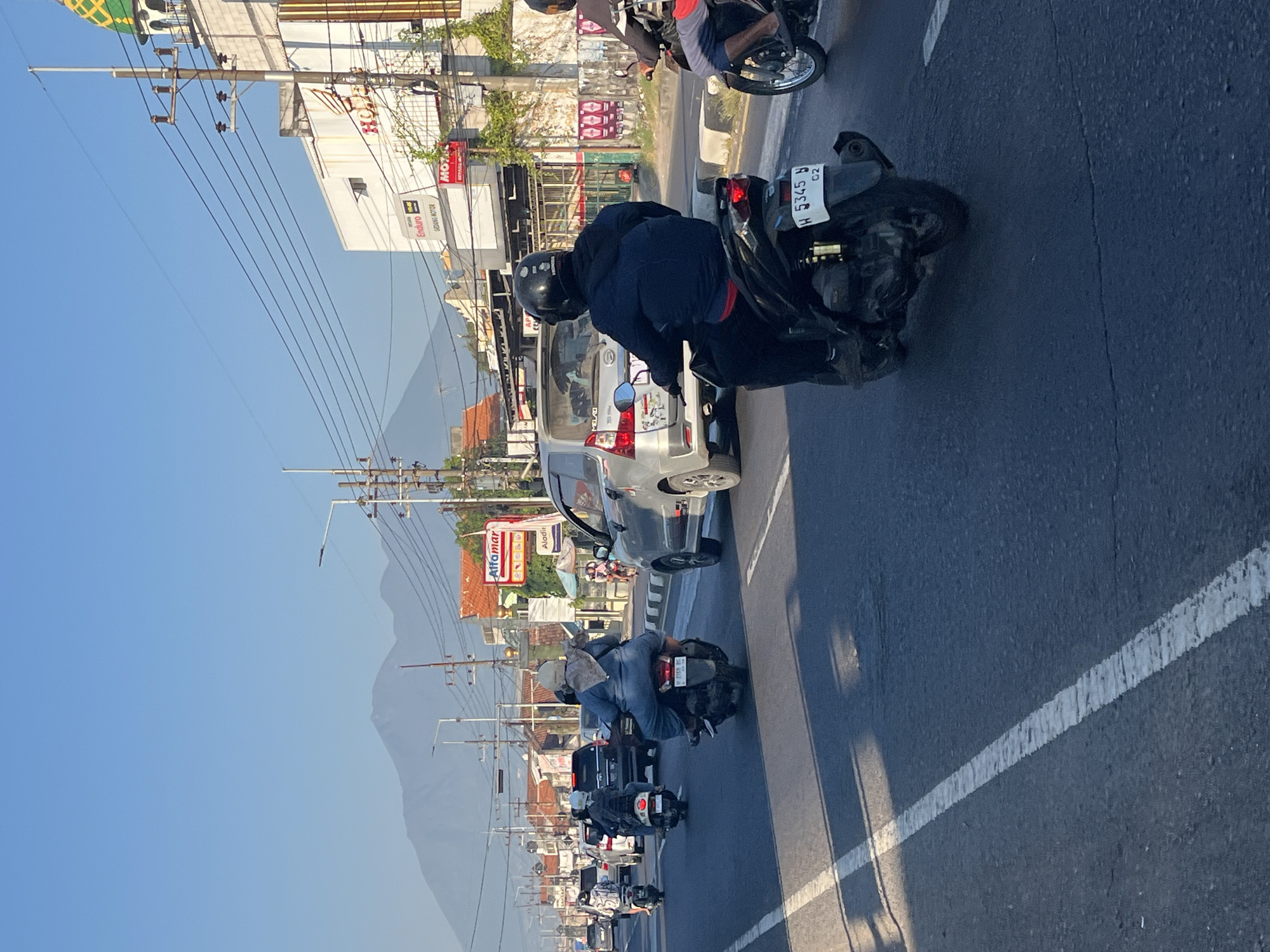 The image captures a street scene in Siring, Indonesia, under a clear blue sky. A road with white lane markings runs through the center, with several vehicles on it. In the foreground, a person rides a dark-colored scooter, wearing a black helmet and a dark blue jacket. The scooter has a white license plate with the number "W 5345 Y". To the left of this scooter, another scooter is visible, with a rider wearing a light brown headscarf and a blue outfit. Further down the road, several other motorcycles and cars are present, some of which are in motion.
On the right side of the street, a white building with a green domed roof stands prominently. A red sign with "Honda" in white letters is visible on this building. Adjacent to it, a smaller red sign displays "MOT" with white lettering and green borders. Another building with an orange roof is seen behind it. In the background, a mountain range looms under the bright sky. Power lines and streetlights are also part of the urban infrastructure. The overall atmosphere suggests a typical day in a bustling Indonesian city.