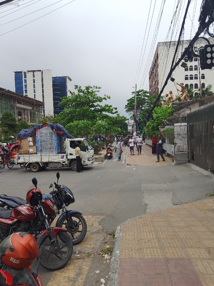 The image shows a street in Dhaka, Bangladesh, at latitude 23.778 and longitude 90.375. Two motorcycles are parked on the right side of the street. A white truck is parked in the middle of the street, facing to the left. A man in a white shirt is standing near the truck. There are people walking on the sidewalk to the right. There is a building to the right with a gate, and a large statue of a man on a horse is visible on the roof. Several buildings are visible in the background. The sky is overcast.