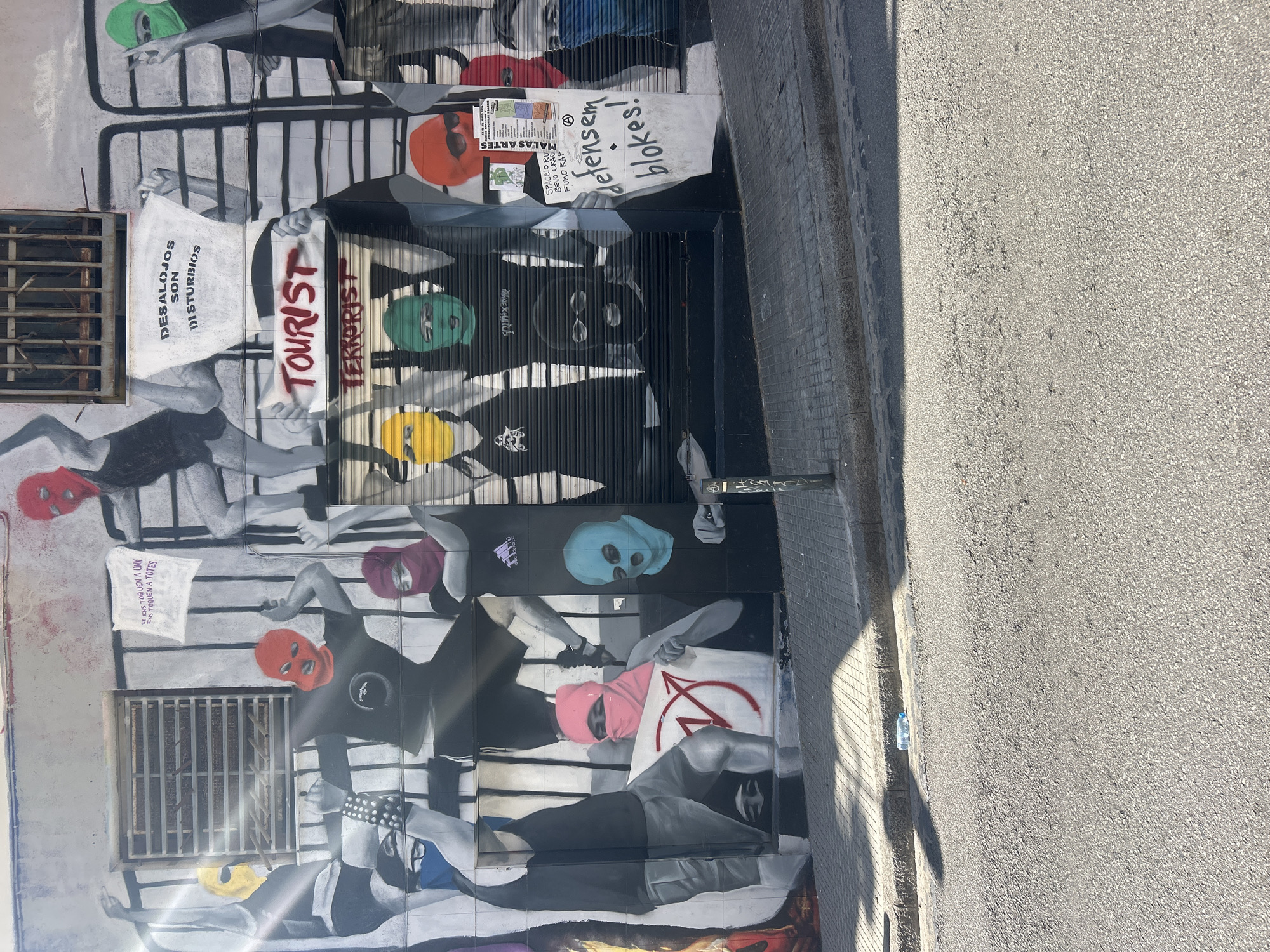 This image captures street art on a wall in Barcelona, Spain. The artwork depicts a series of figures wearing colorful ski masks and dark clothing, some holding signs with various political messages. The messages are written in different languages, including "DESALOJOS SON DISTURBIOS" (Evictions are disturbances), "TOURIST TERRORIST", "etensem blokes!", and "ENS TOQUEN A UNY ENS TOQUEN A TOTES". The wall features architectural details like barred windows. The scene includes a section of a street and sidewalk.