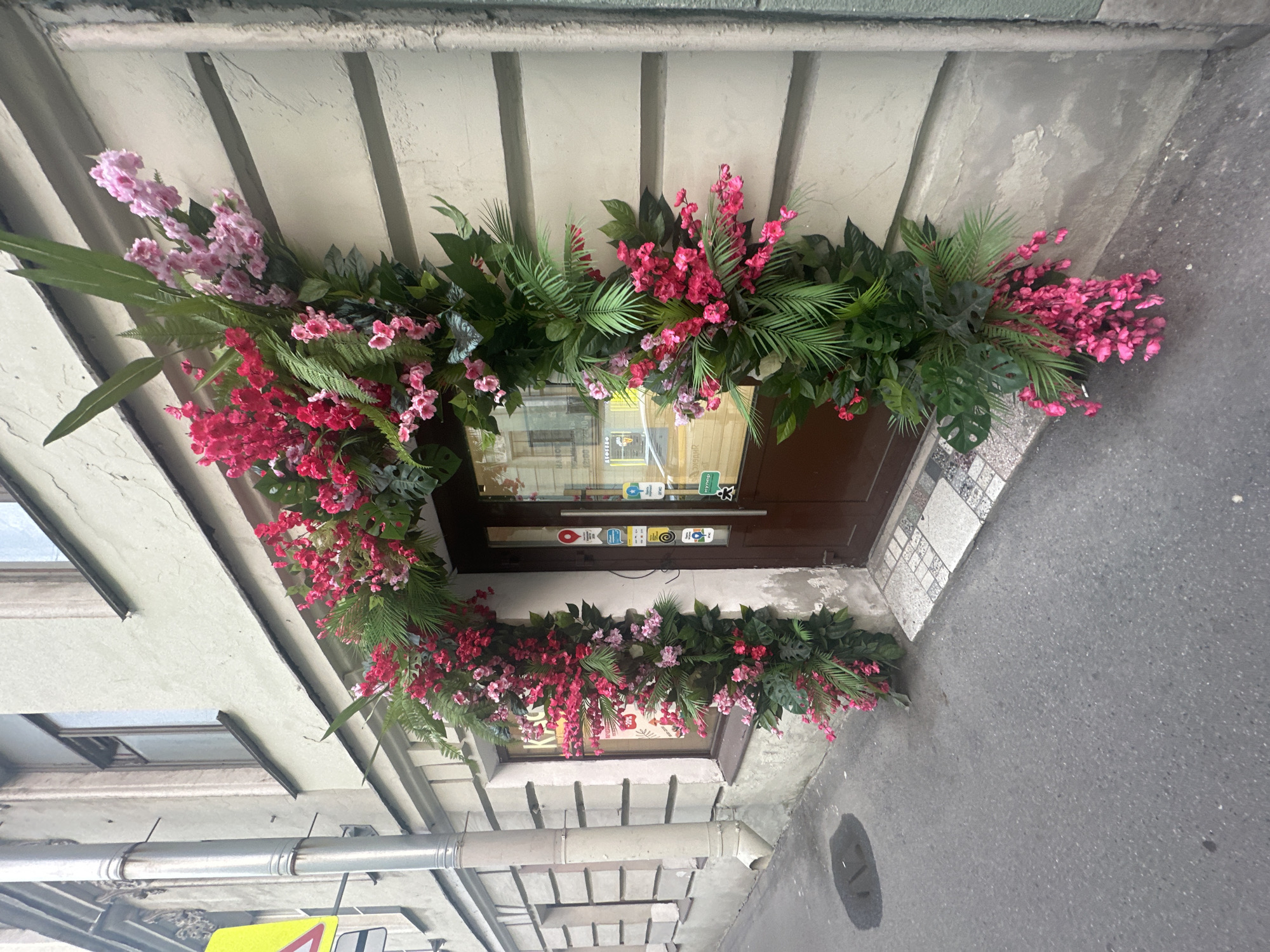 A building entrance is elaborately decorated with an arrangement of pink and red flowers intertwined with greenery. The floral arrangement spans the top and sides of the doorway, creating a colorful frame. The door itself is dark brown and features signage and stickers. The building's facade is light-colored with a horizontally-grooved pattern. The scene is captured from a low angle, looking upwards towards the entrance, and the sidewalk and part of the street are visible at the bottom right. The photograph was taken in Saint Petersburg, Russia.